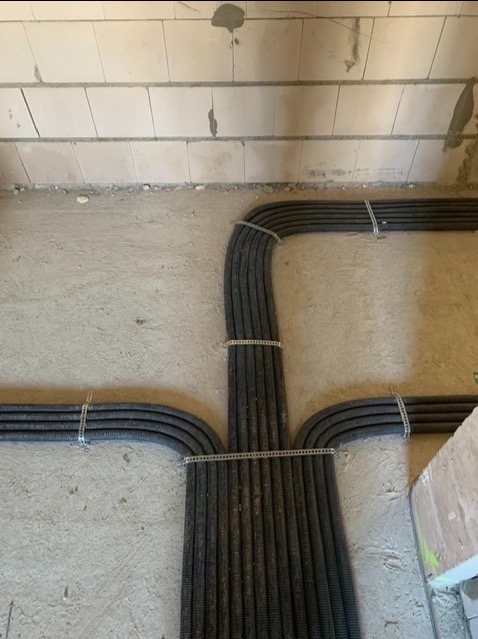 Układ czarnych, karbowanych rur instalacyjnych przytwierdzonych do surowej ściany za pomocą metalowych uchwytów. Rury rozchodzą się w trzech kierunkach, tworząc symetryczny wzór na tle szarego...