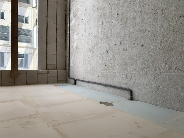 Szara rura instalacji elektrycznej poprowadzona wzdłuż surowej ściany w stanie deweloperskim, widok na budynek z oknami w tle przez niezabezpieczony otwór okienny.