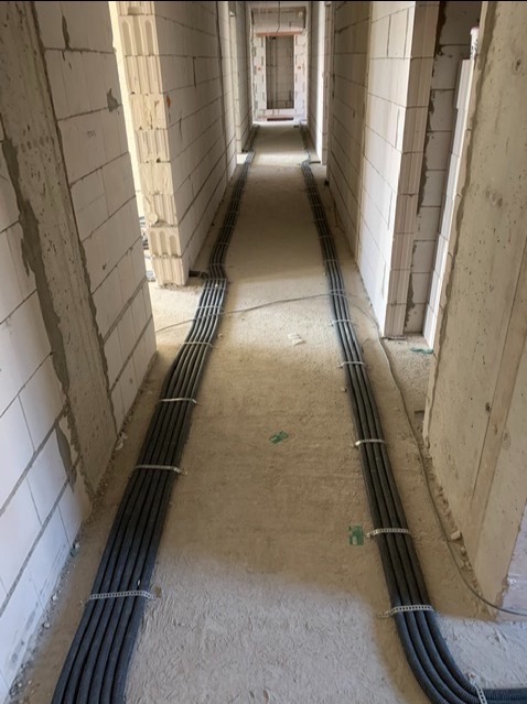 Kable elektryczne w peszlach ułożone równolegle wzdłuż korytarza w surowym stanie, przymocowane metalowymi uchwytami do podłoża, widok w perspektywie.