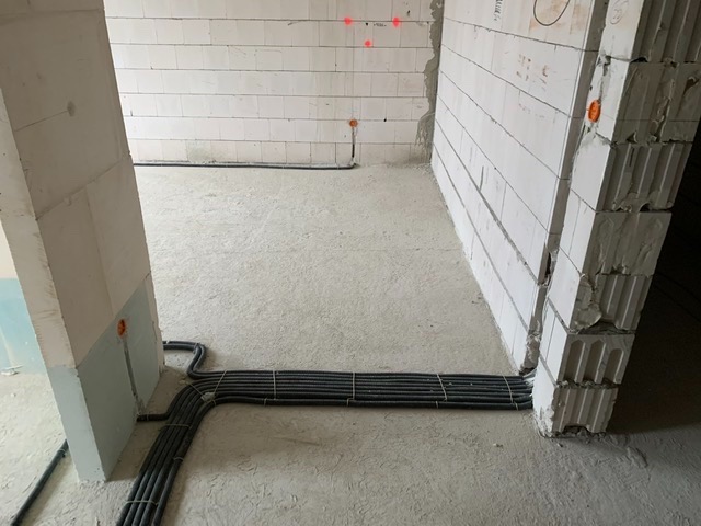 Wiązka czarnych kabli elektrycznych ułożona na surowym betonowym podłożu w trakcie prac instalacyjnych w nowo budowanym pomieszczeniu z białych bloczków, widoczne pomarańczowe puszki elektryczne...