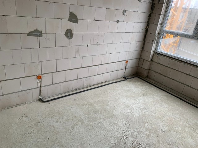 Surowy stan wnętrza z widoczną instalacją elektryczną: czarny przewód biegnie wzdłuż ściany z białego bloczka, zakończony pomarańczową puszką. W tle widoczne okno i fragment żurawia budowlanego.