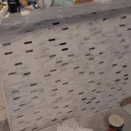 Ściana baru wyłożona mozaiką z białych i szarych prostokątnych płytek, widoczne narzędzia glazurnicze, w tym szlifierka kątowa i przymiar, oraz materiały budowlane na blacie, na którym stoi puszka...