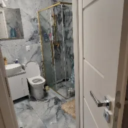 Wykończona łazienka z marmurowymi płytkami, złotą kabiną prysznicową i białymi meblami, widok zza uchylonych drzwi.