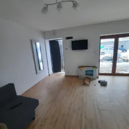 Salon po remoncie z drewnopodobnymi panelami podłogowymi, widoczna kanapa, telewizor na ścianie i karton po telewizorze na podłodze.