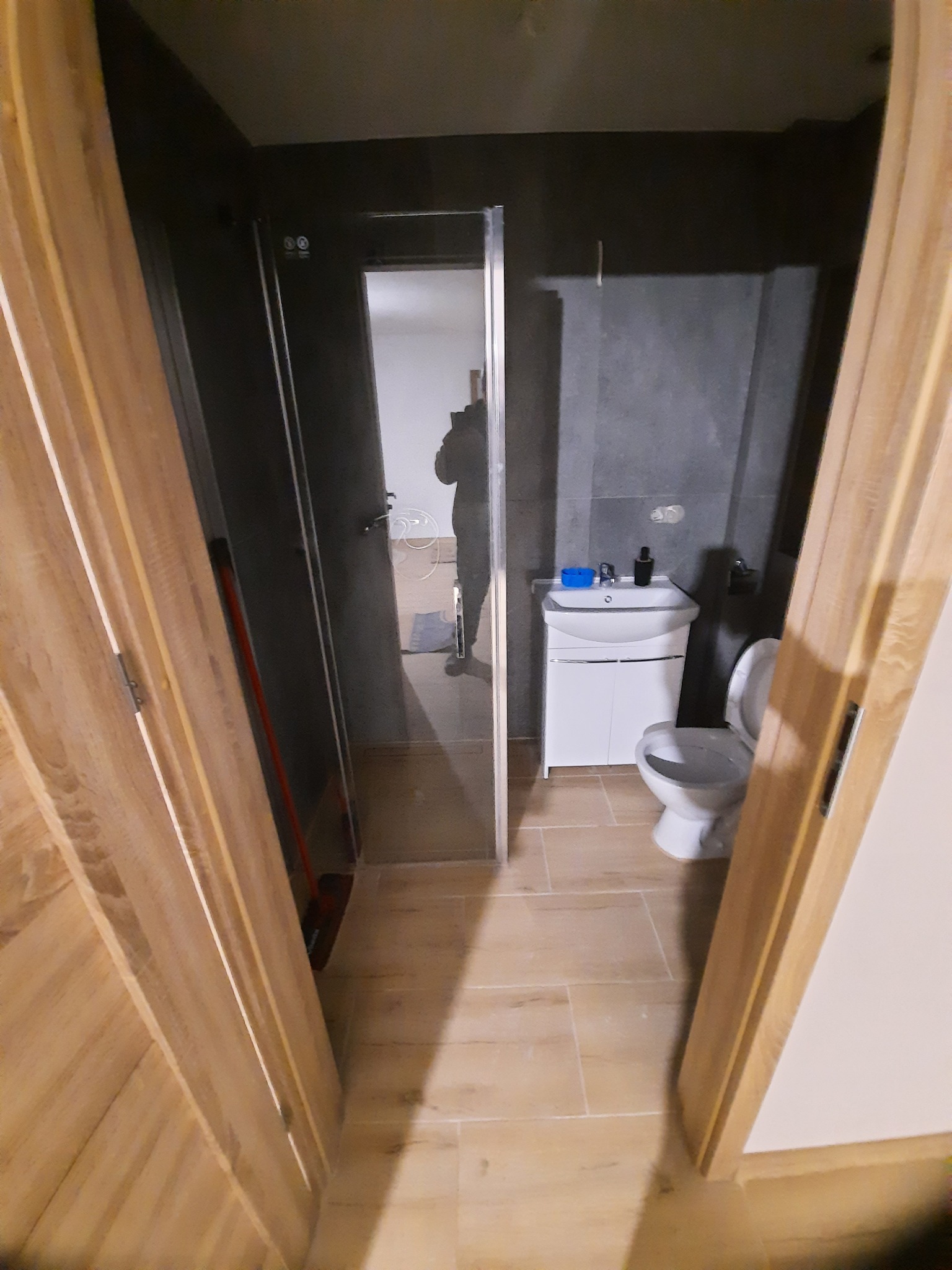 Widok na odnowioną łazienkę z kabiną prysznicową, umywalką na szafce i toaletą, wykończoną płytkami w odcieniach beżu i szarości, widziany przez otwarte drewniane drzwi.