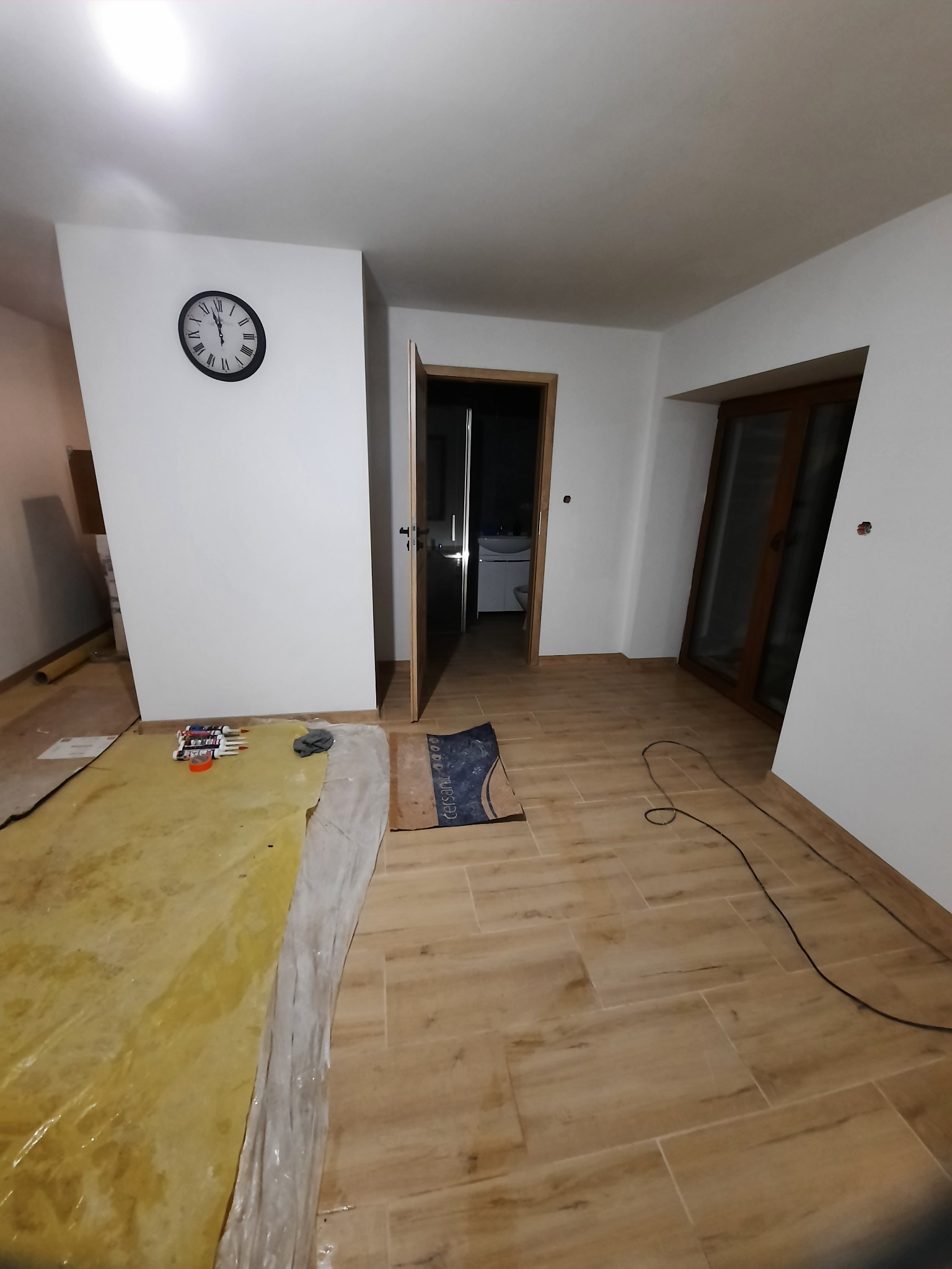 Jasne płytki podłogowe imitujące drewno w pokoju podczas remontu, zabezpieczona folią część podłogi, widoczne narzędzia i otwarte drzwi do łazienki.