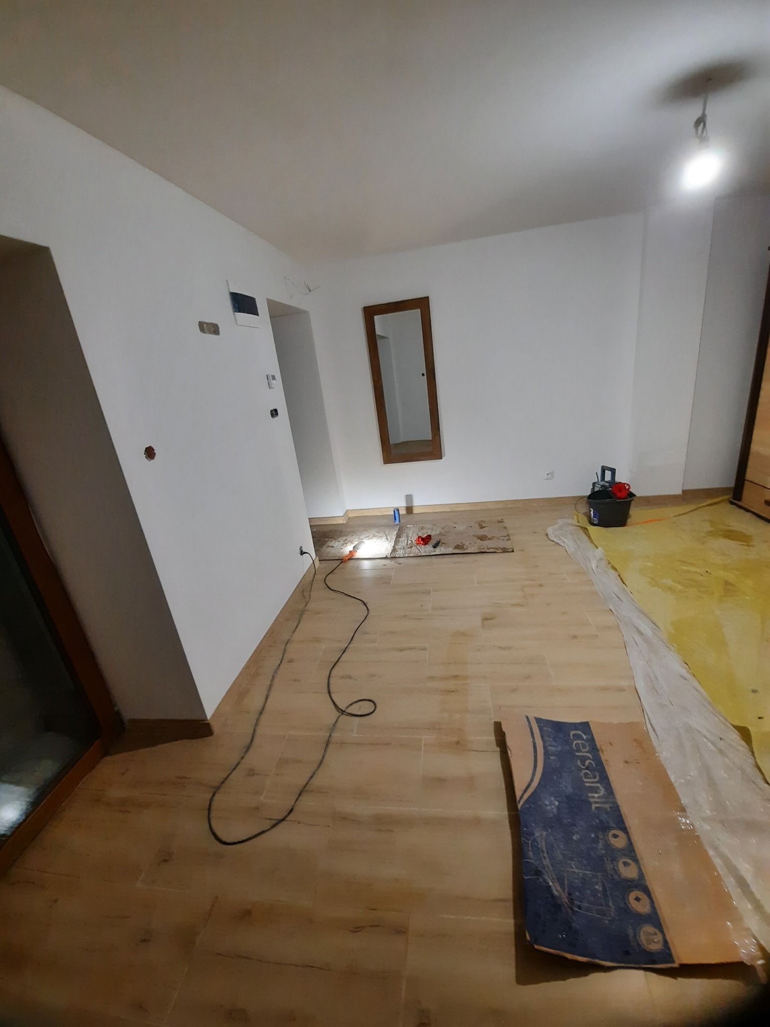Pokój w trakcie remontu: podłoga wyłożona panelami imitującymi drewno, ściany przygotowane do malowania z widocznymi otworami na gniazdka, karton po panelach podłogowych i narzędzia malarskie...