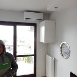 Paweł Romańczuk - Biała jednostka klimatyzacji zamontowana nad oknem balkonowym w nowoczesnym salonie, z widoczną częścią osoby w bluzie z kapturem i elementami wystroju wnętrza, takimi jak białe szafki wiszące...