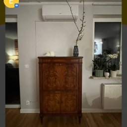 Paweł Romańczuk - Wnętrze pokoju z zamontowaną klimatyzacją ścienną nad stylową, drewnianą komodą. Na komodzie wazon z gałązkami, obok okno z roślinami doniczkowymi i grzejnikiem.