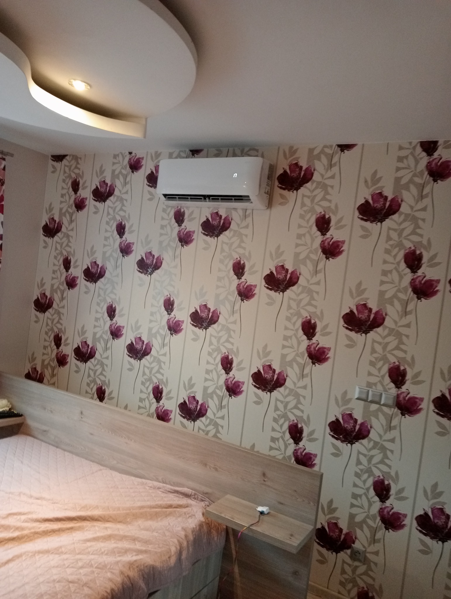Biała jednostka klimatyzacyjna zamontowana na ścianie w sypialni z tapetą w kwiaty, nad łóżkiem z drewnianym zagłówkiem.
