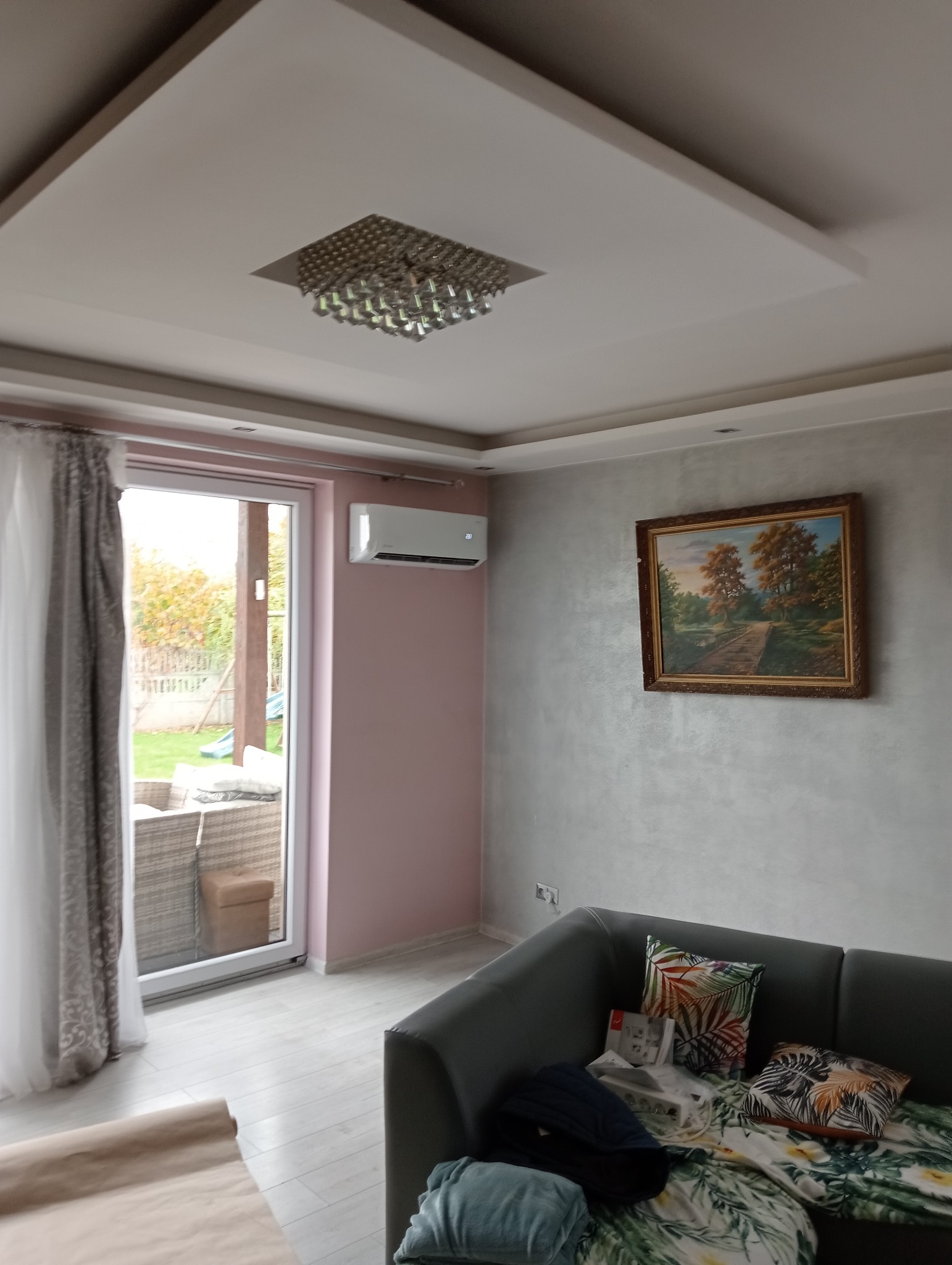 Wnętrze salonu z zamontowaną jednostką klimatyzacyjną na ścianie, widok na okno balkonowe, szary narożnik z poduszkami dekoracyjnymi i obraz na ścianie.