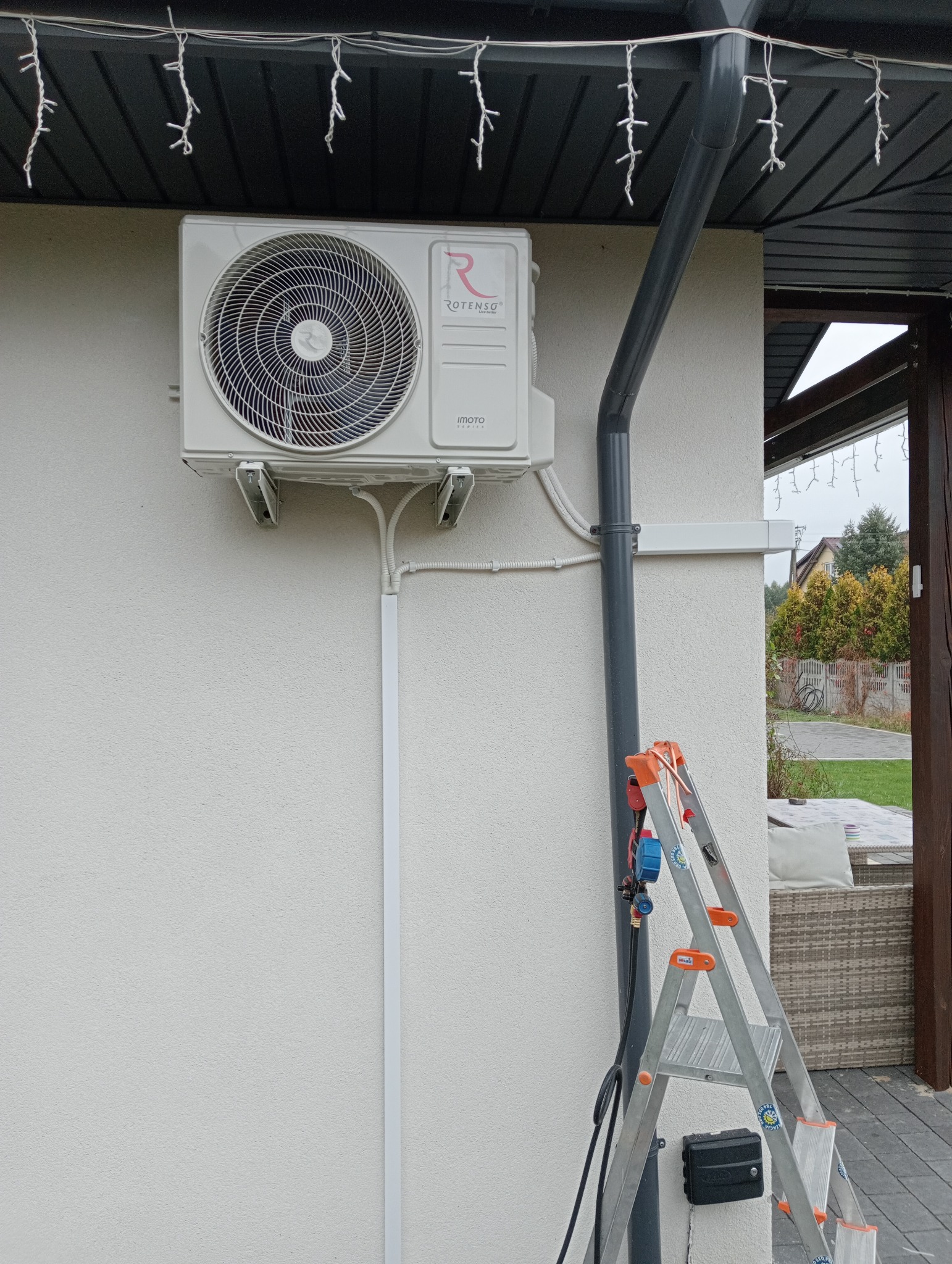 Zamontowana jednostka zewnętrzna klimatyzacji Rotenso na elewacji domu, widoczna drabina oparta o ścianę i rurę spustową, białe listwy maskujące instalację elektryczną i rury chłodnicze.