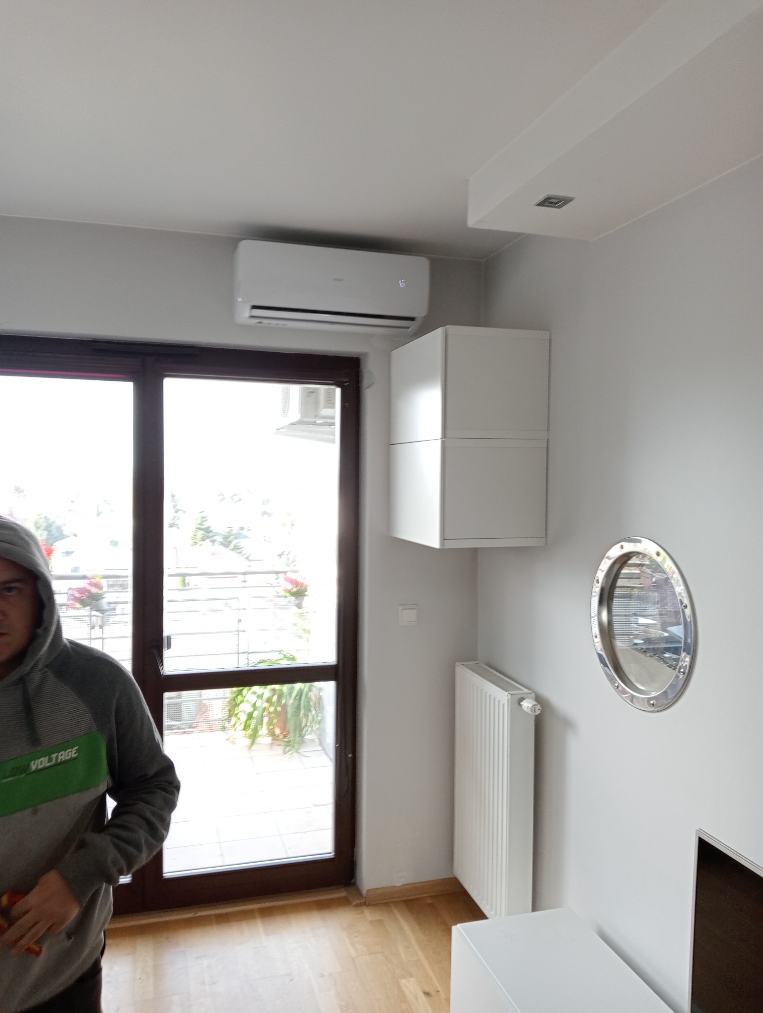 Biała jednostka klimatyzacji zamontowana nad oknem balkonowym w nowoczesnym salonie, z widoczną częścią osoby w bluzie z kapturem i elementami wystroju wnętrza, takimi jak białe szafki wiszące...