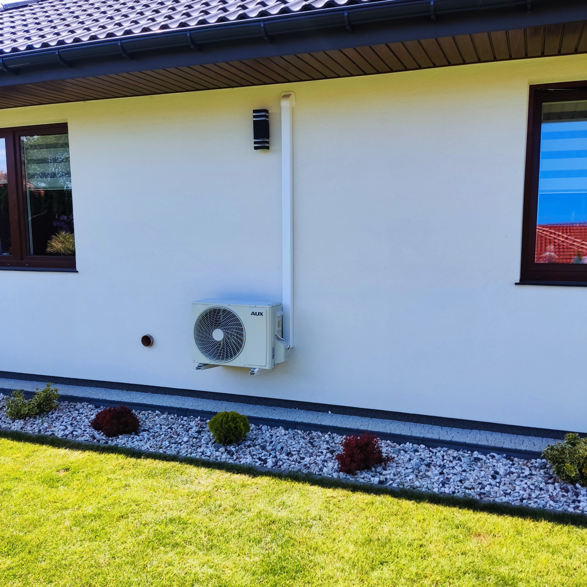 Zewnętrzna jednostka klimatyzatora AUX zamontowana na białej ścianie domu, z widoczną instalacją i otoczeniem w postaci trawnika i ozdobnych krzewów.