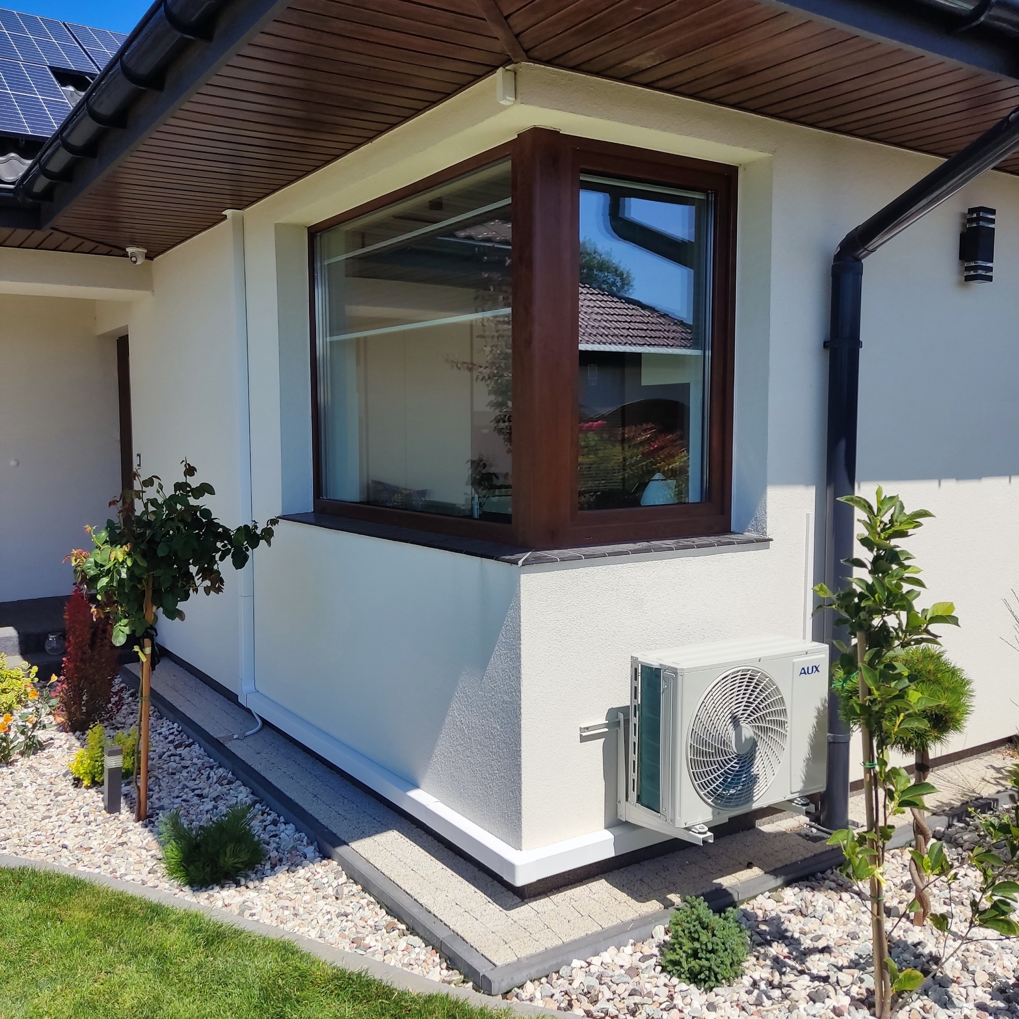 Zewnętrzna jednostka klimatyzacji AUX zamontowana na elewacji domu z brązową stolarką okienną, otoczona ozdobnymi krzewami i kamieniami.