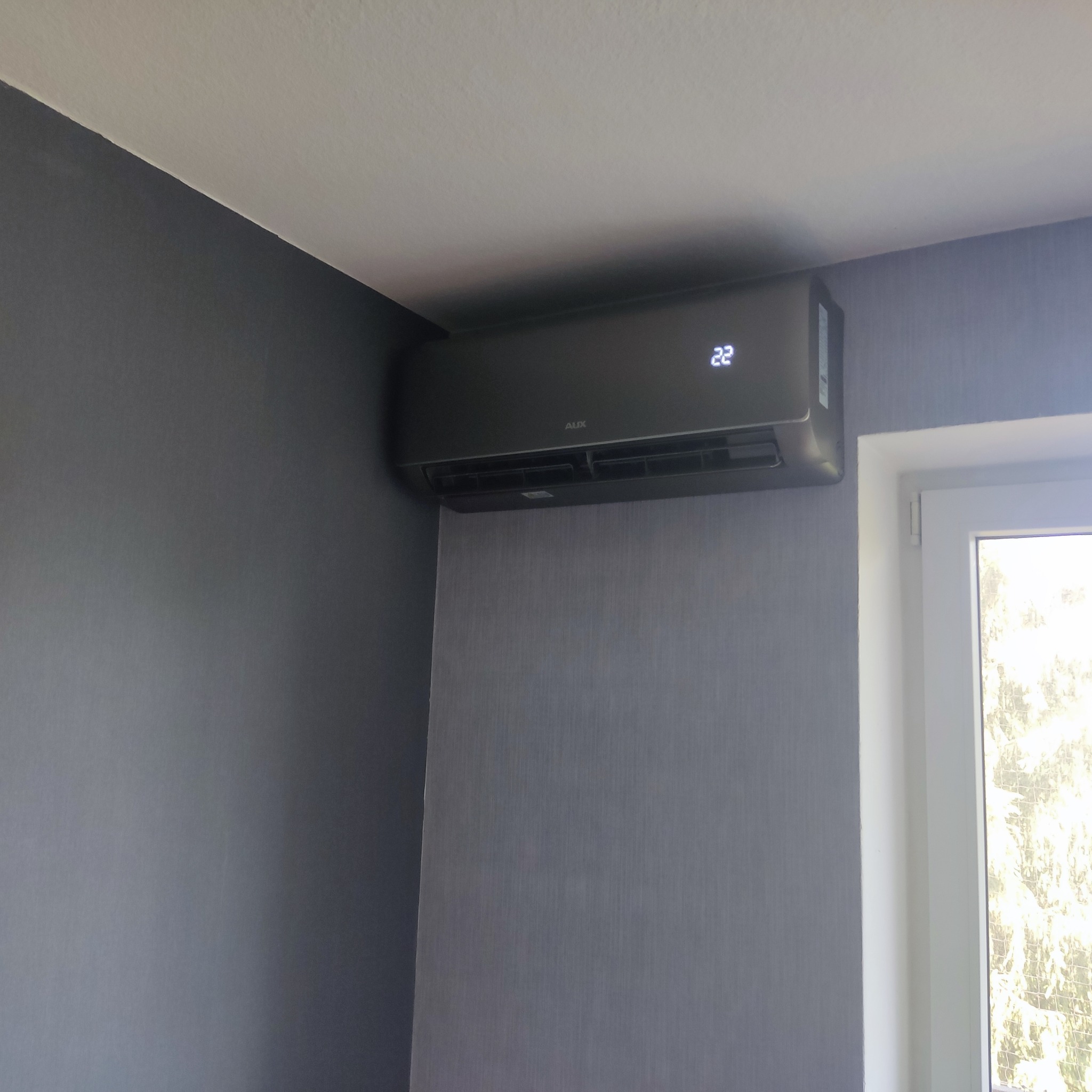 Czarny klimatyzator marki AUX zamontowany na ścianie w szarym pokoju, obok okna.