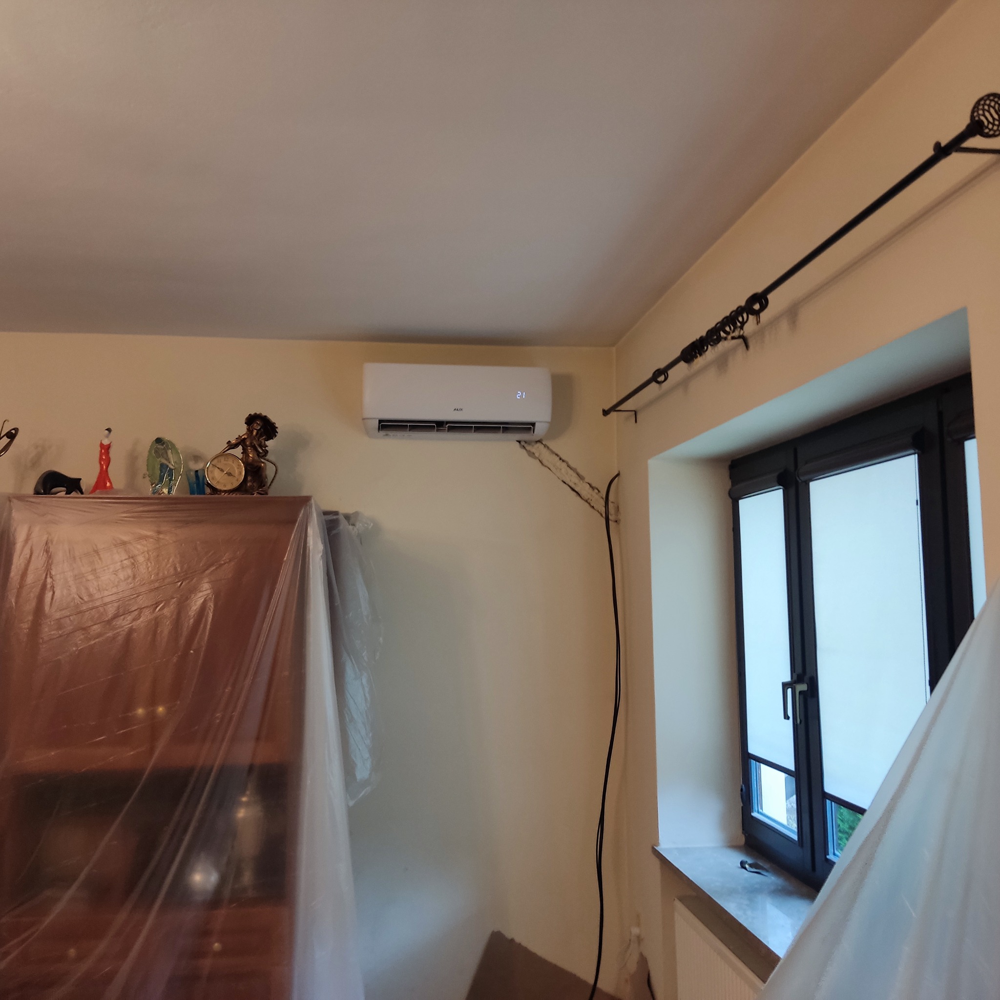 Biały klimatyzator AUX zamontowany na ścianie w pokoju, zabezpieczone meble folią ochronną podczas instalacji, widoczne przewody elektryczne.