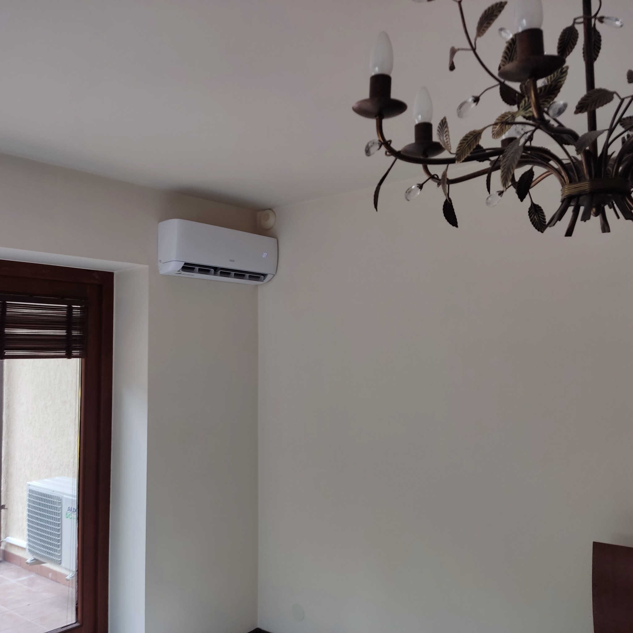 Biały klimatyzator ścienny z wyświetlaczem temperatury w jasnym pomieszczeniu z dekoracyjnym żyrandolem i oknem balkonowym.