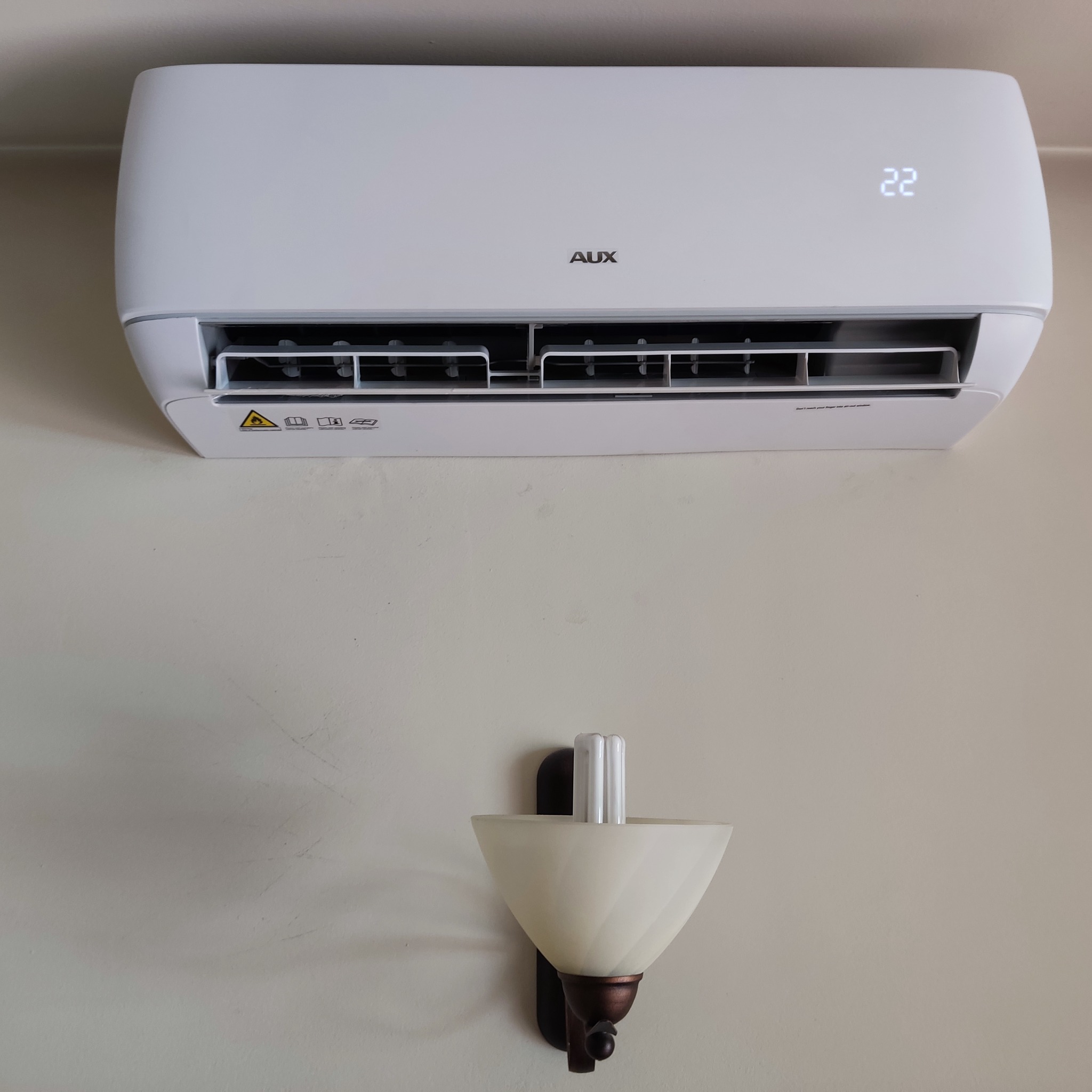 Biały klimatyzator marki AUX zamontowany na ścianie, z wyświetlaczem wskazującym temperaturę 22 stopnie Celsjusza, pod nim kinkiet z energooszczędną żarówką.