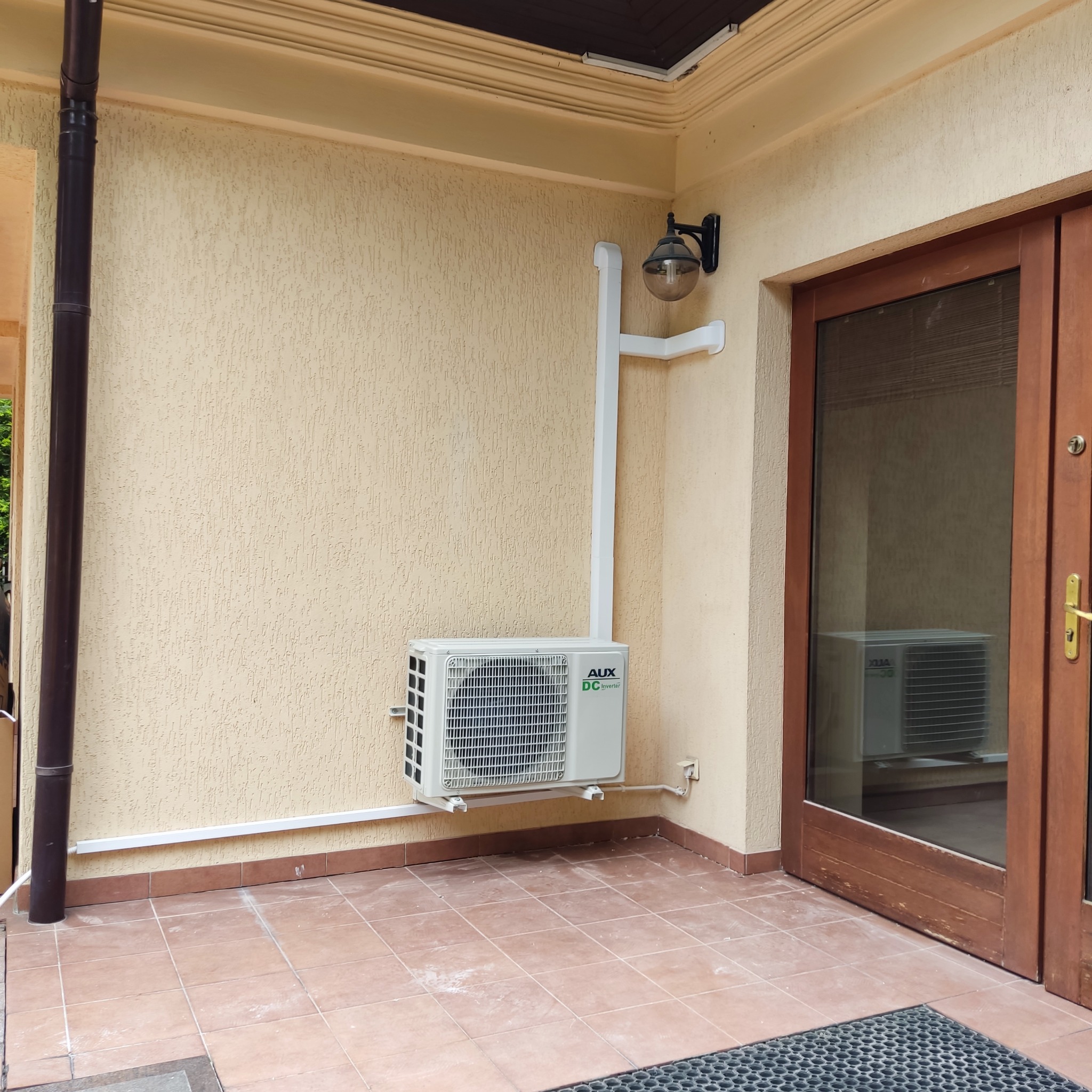 Zewnętrzna jednostka klimatyzacji AUX DC Inverter zamontowana na ścianie budynku z elewacją w kolorze kremowym, widoczny fragment brązowych drzwi z szybą, w której odbija się kolejna jednostka...