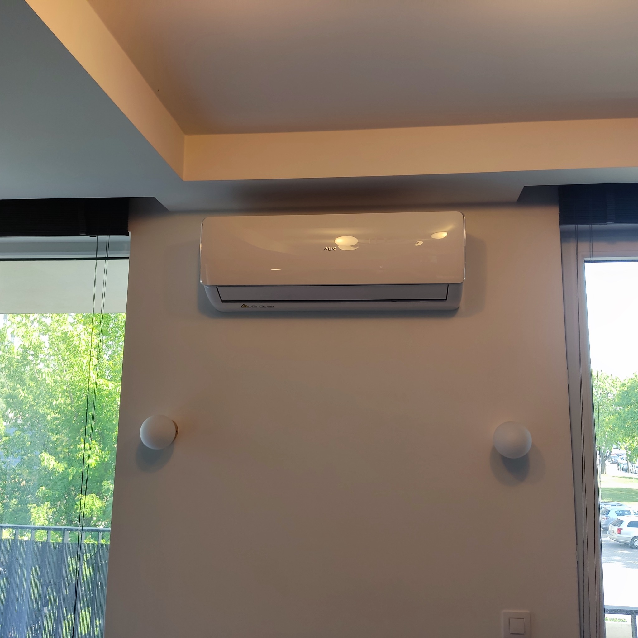 Biały klimatyzator marki AUX zamontowany na ścianie w jasnym pomieszczeniu z widokiem na zieleń za oknem.