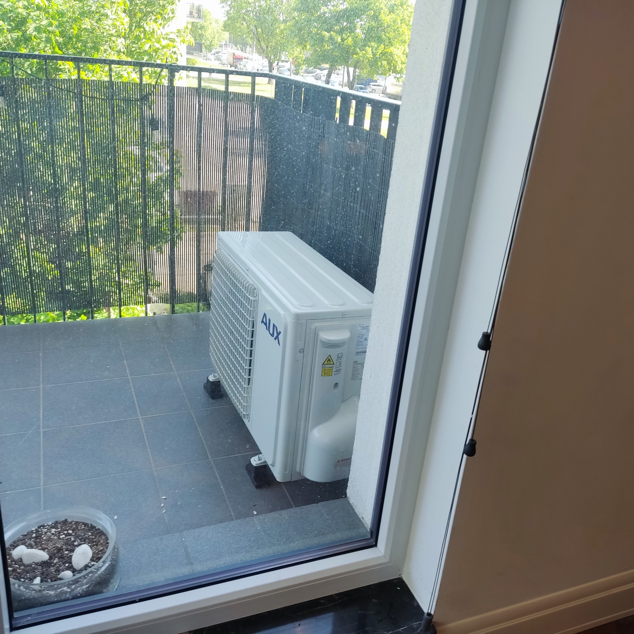 Kompaktowa jednostka zewnętrzna klimatyzacji marki AUX, ustawiona na balkonie z szarymi płytkami, widoczna przez okno. W tle znajduje się metalowa balustrada i drzewa.