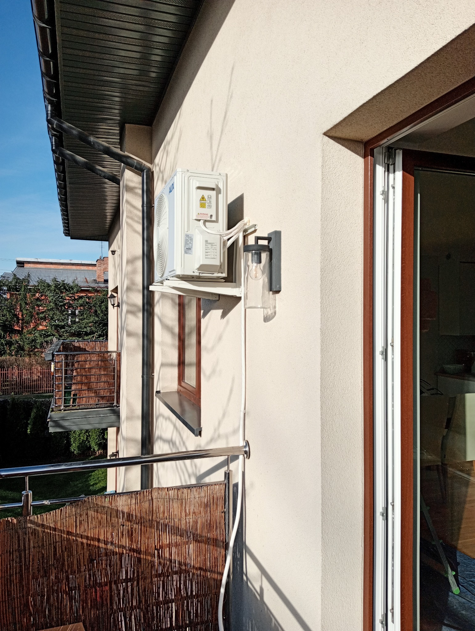 Zewnętrzna jednostka klimatyzacji marki AUX zamontowana na balkonie domu jednorodzinnego, widoczna lampa ścienna i fragment balustrady z matą bambusową.