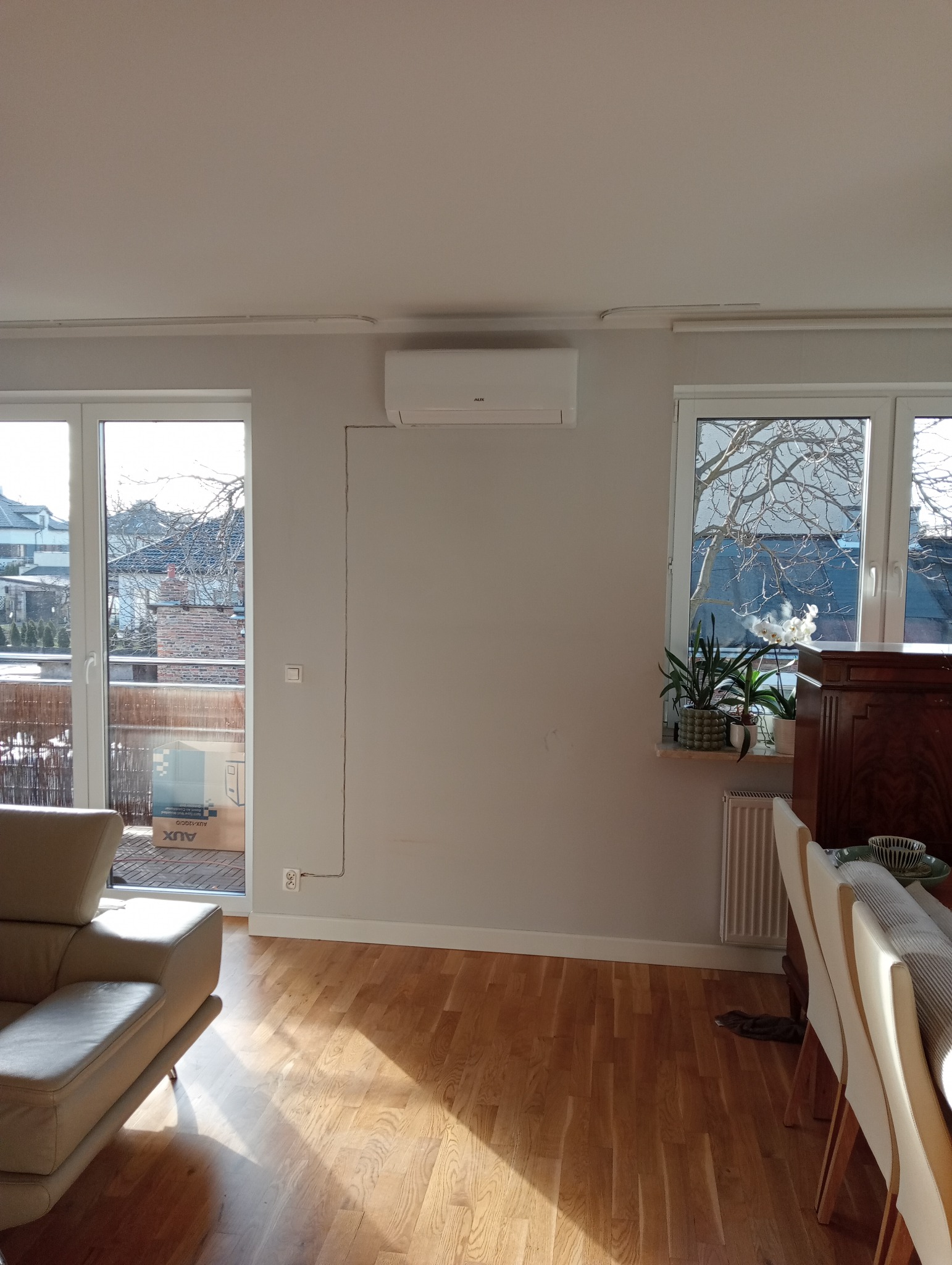 Biała jednostka klimatyzacyjna AUX zamontowana na ścianie w jasnym salonie, z widocznymi przewodami i skrzynką AUX na balkonie, oświetlonym popołudniowym słońcem.