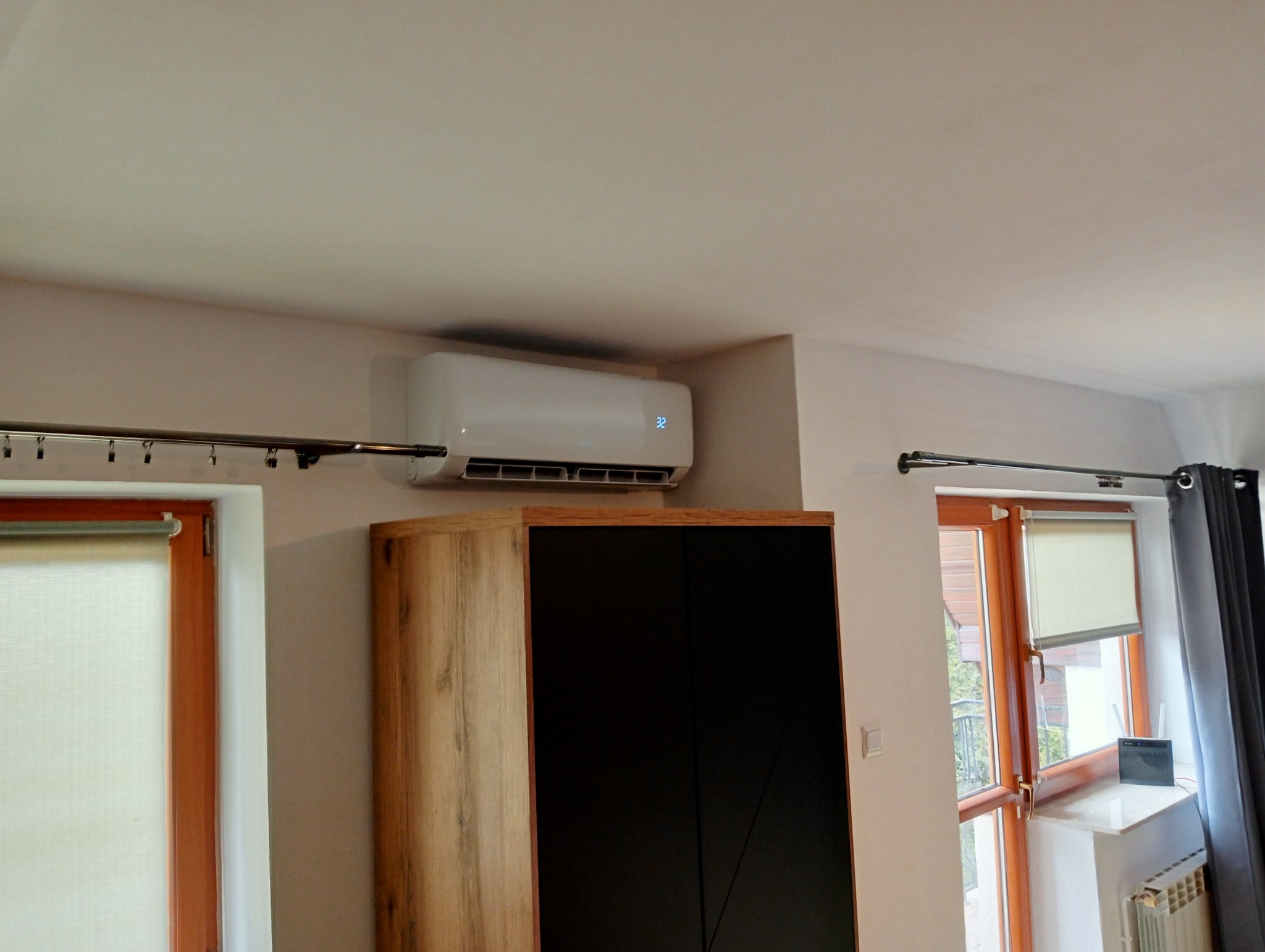 Biały klimatyzator ścienny z wyświetlaczem temperatury 32 stopnie Celsjusza, zamontowany nad szafą w pokoju z drewnianymi oknami i roletami.