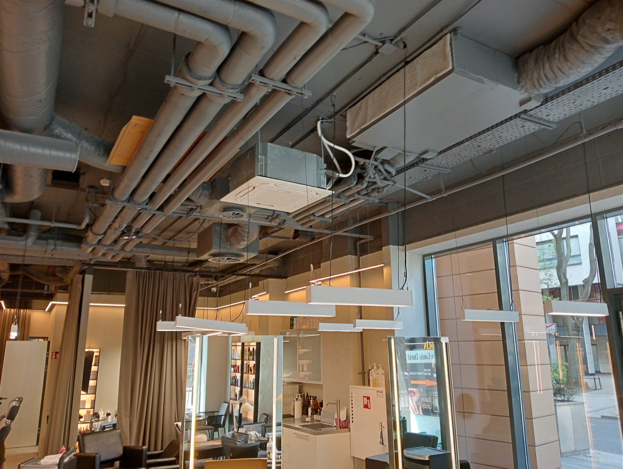 Sufit w salonie fryzjerskim z widoczną instalacją wentylacyjną i klimatyzacyjną, rurami, kablami oraz oświetleniem.