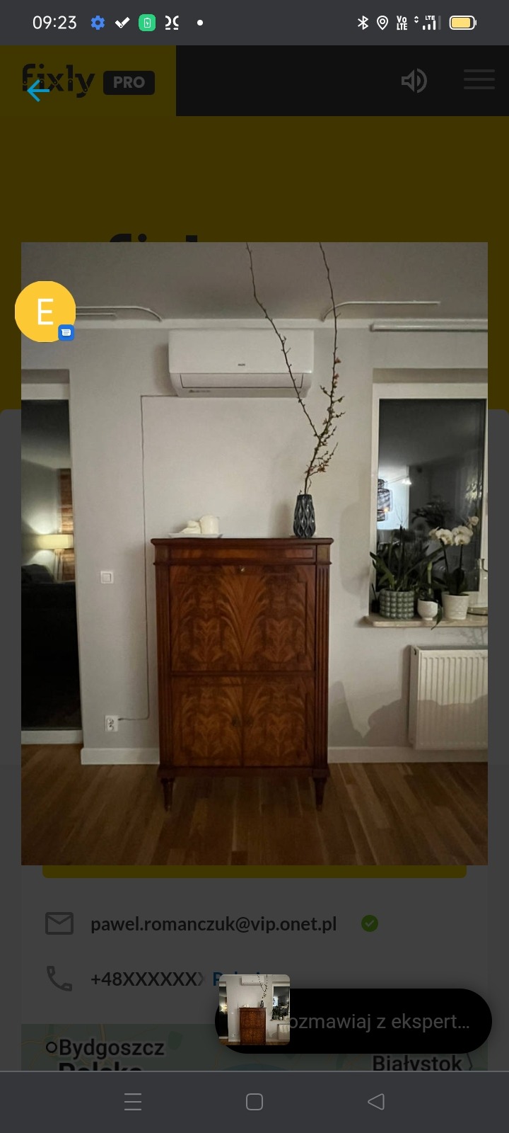 Wnętrze pokoju z zamontowaną klimatyzacją ścienną nad stylową, drewnianą komodą. Na komodzie wazon z gałązkami, obok okno z roślinami doniczkowymi i grzejnikiem.