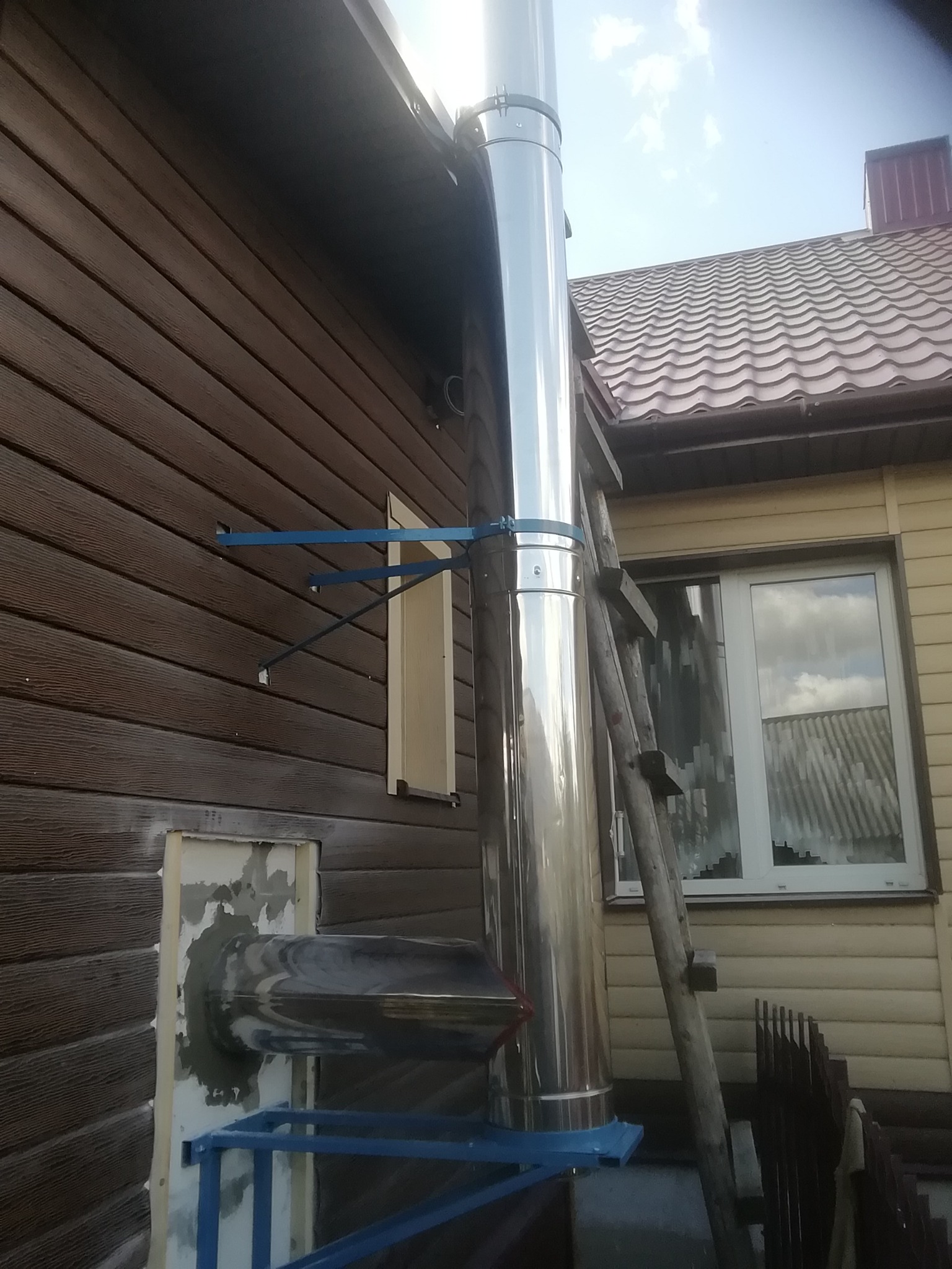 Montaż stalowego komina zewnętrznego w Warszawie, widoczne mocowania do brązowej elewacji oraz drabina oparta o ścianę budynku.
