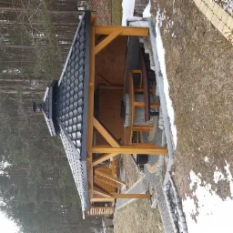 Zadaszona altana z drewnianą konstrukcją, czarnym dachem z kominem i brukowaną podłogą, widoczny stół i ławki wewnątrz, obok drzewa i trawnik z resztkami śniegu.