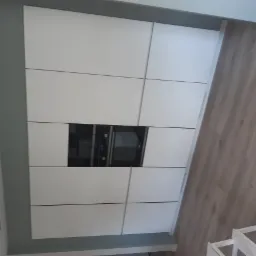 Nowoczesna zabudowa kuchenna z białymi frontami i wbudowanym czarnym piekarnikiem oraz mikrofalówką, widok z góry na drewnianą podłogę.