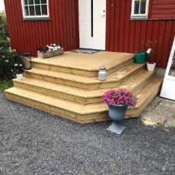 Nowe, drewniane schody wejściowe prowadzące do domu z czerwonymi ścianami, ozdobione kwiatami w donicach i skrzynkach.