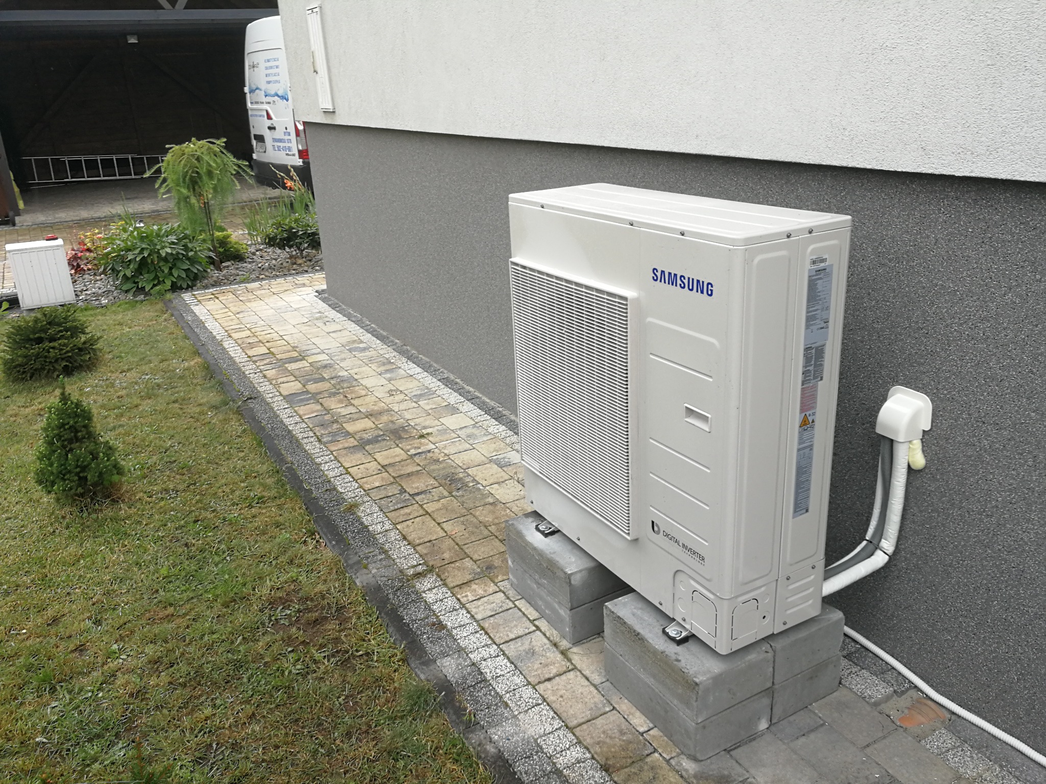 Biała, zewnętrzna jednostka klimatyzacji Samsung Digital Inverter na betonowych bloczkach przy ścianie budynku z widocznymi przewodami i kostką brukową.