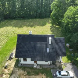 Widok z góry na dom jednorodzinny z ciemną dachówką i zamontowanymi panelami słonecznymi; obok domu zaparkowany biały samochód, a w tle zielony teren z drzewami.