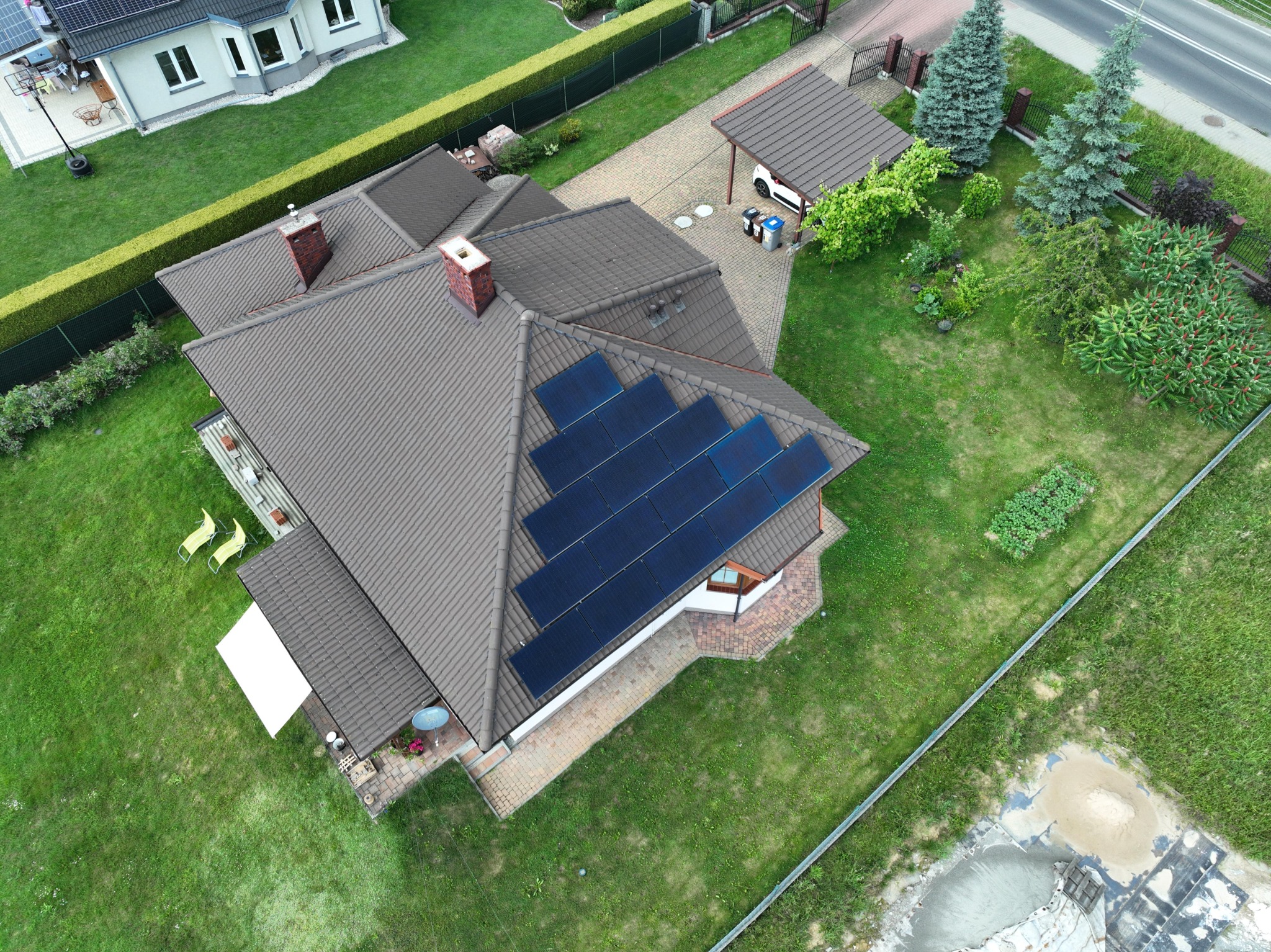 Widok z drona na dom jednorodzinny z brązowym dachem i panelami słonecznymi, otoczony zielonym trawnikiem i ogrodzeniem z tujami. Na podjeździe widoczny biały samochód i kosze na śmieci.