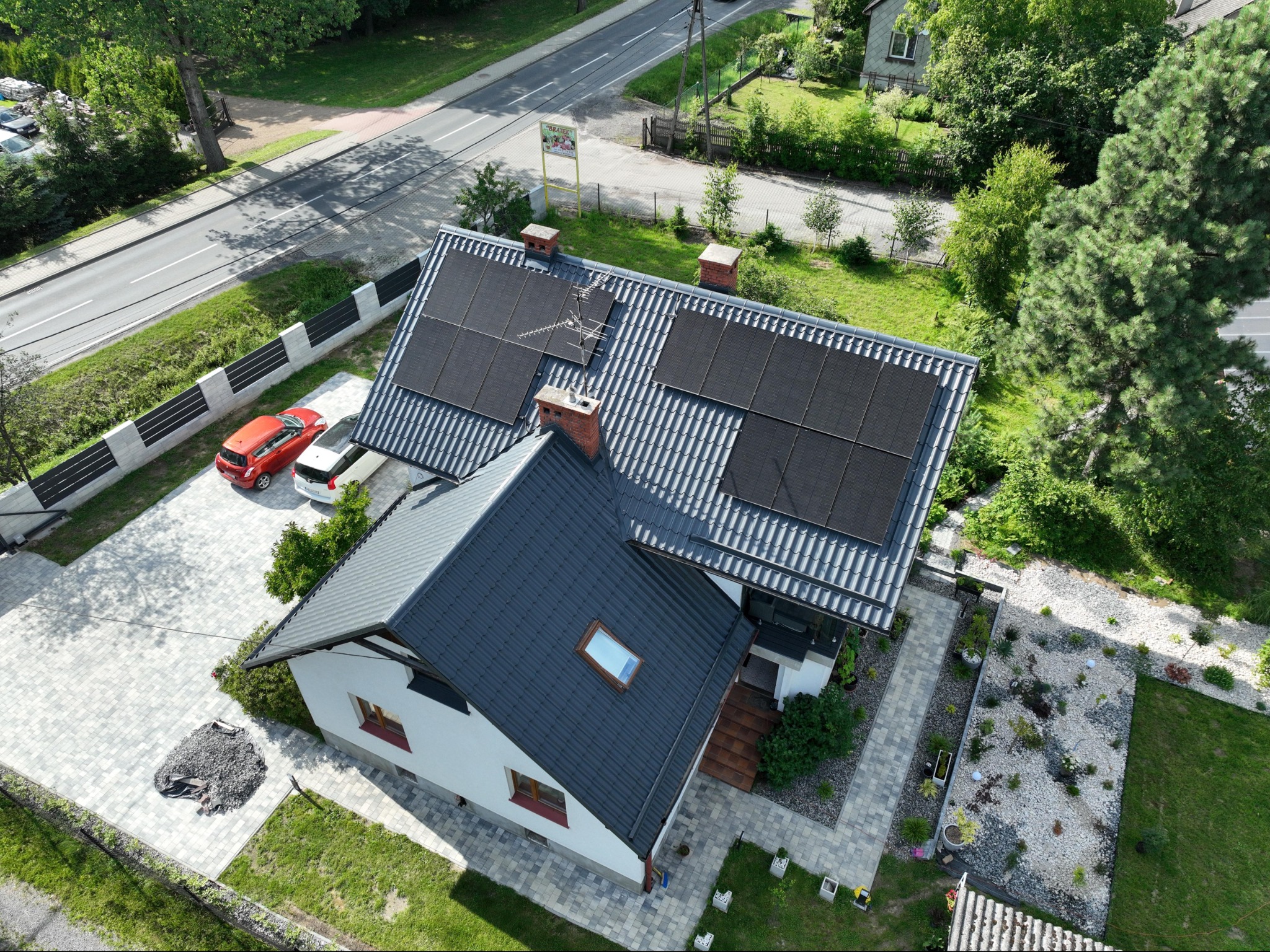 Widok z góry na dom z zainstalowanymi panelami słonecznymi na dachu, antena telewizyjna, czerwony samochód i szara kostka brukowa na podjeździe.