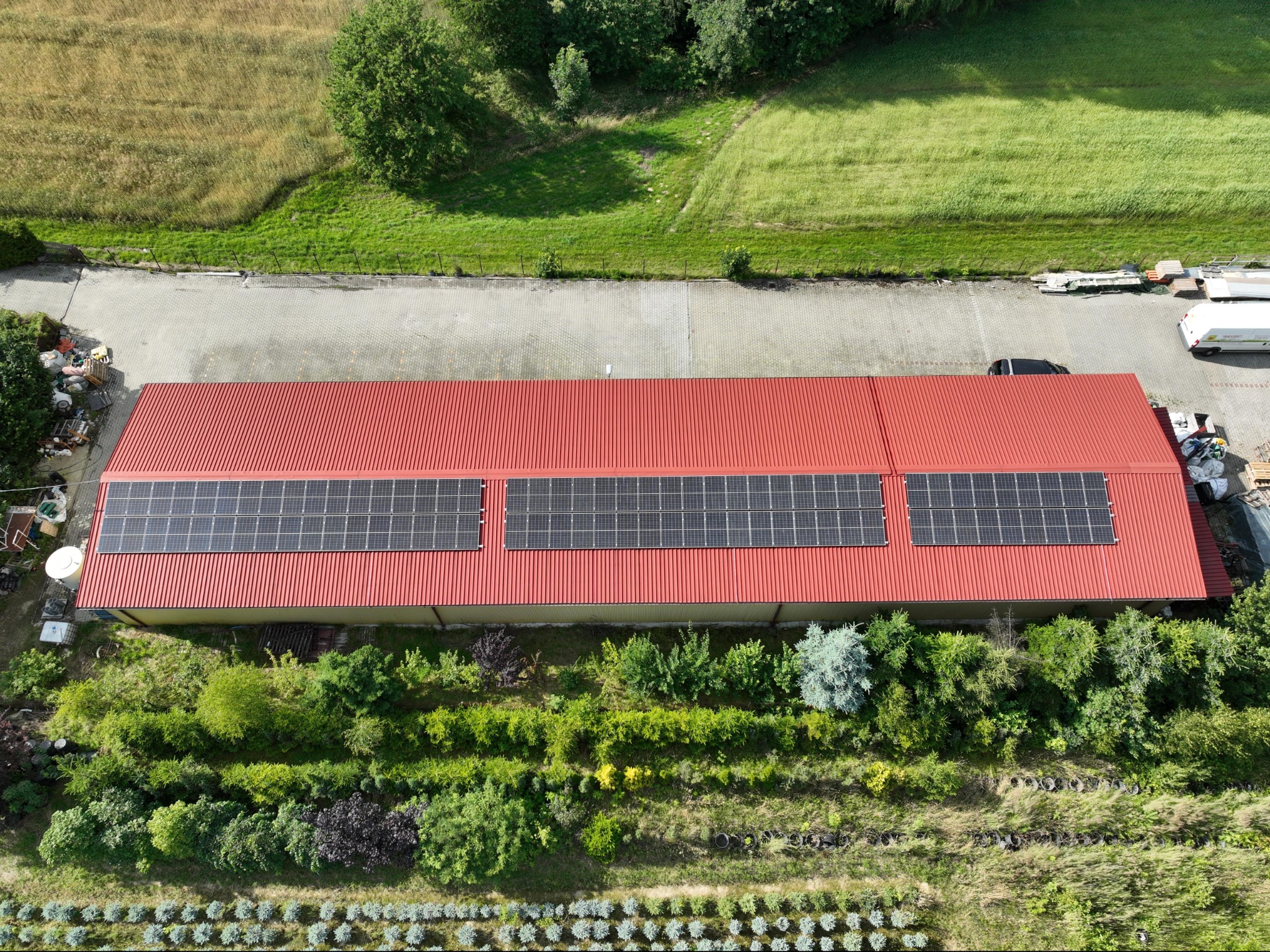 Widok z góry na czerwony dach budynku z trzema panelami słonecznymi, otoczony zielenią, z polami i drzewami w tle.