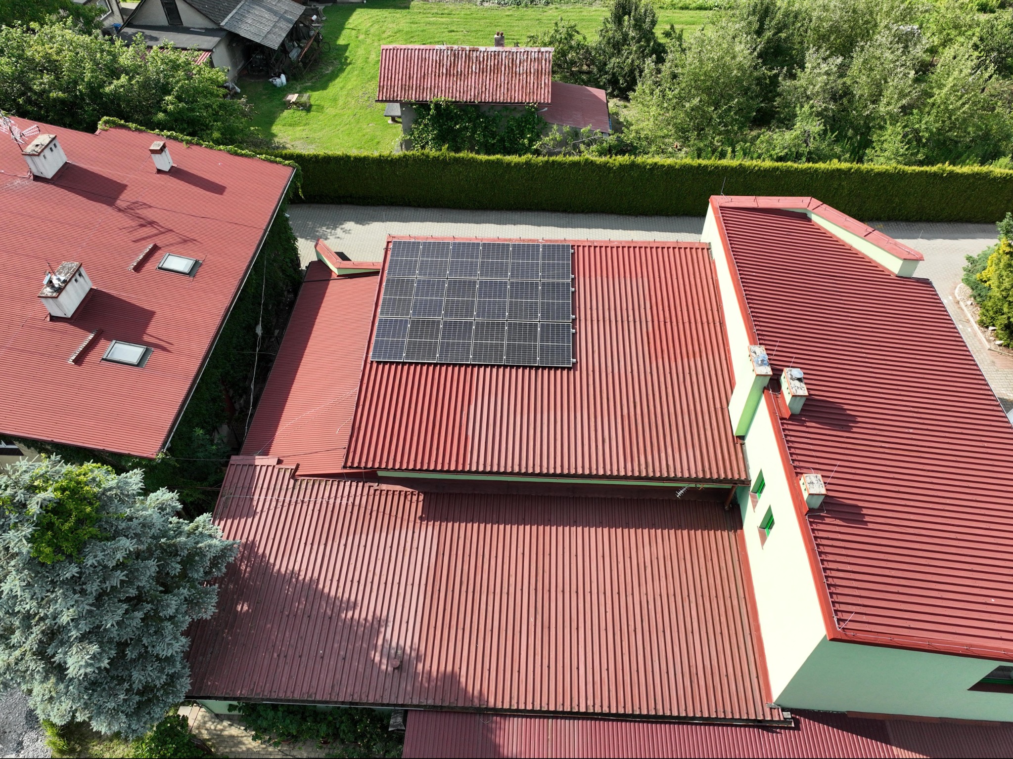 Widok z góry na panele fotowoltaiczne zamontowane na czerwonym, blaszany dachu budynku mieszkalnego w otoczeniu zieleni.