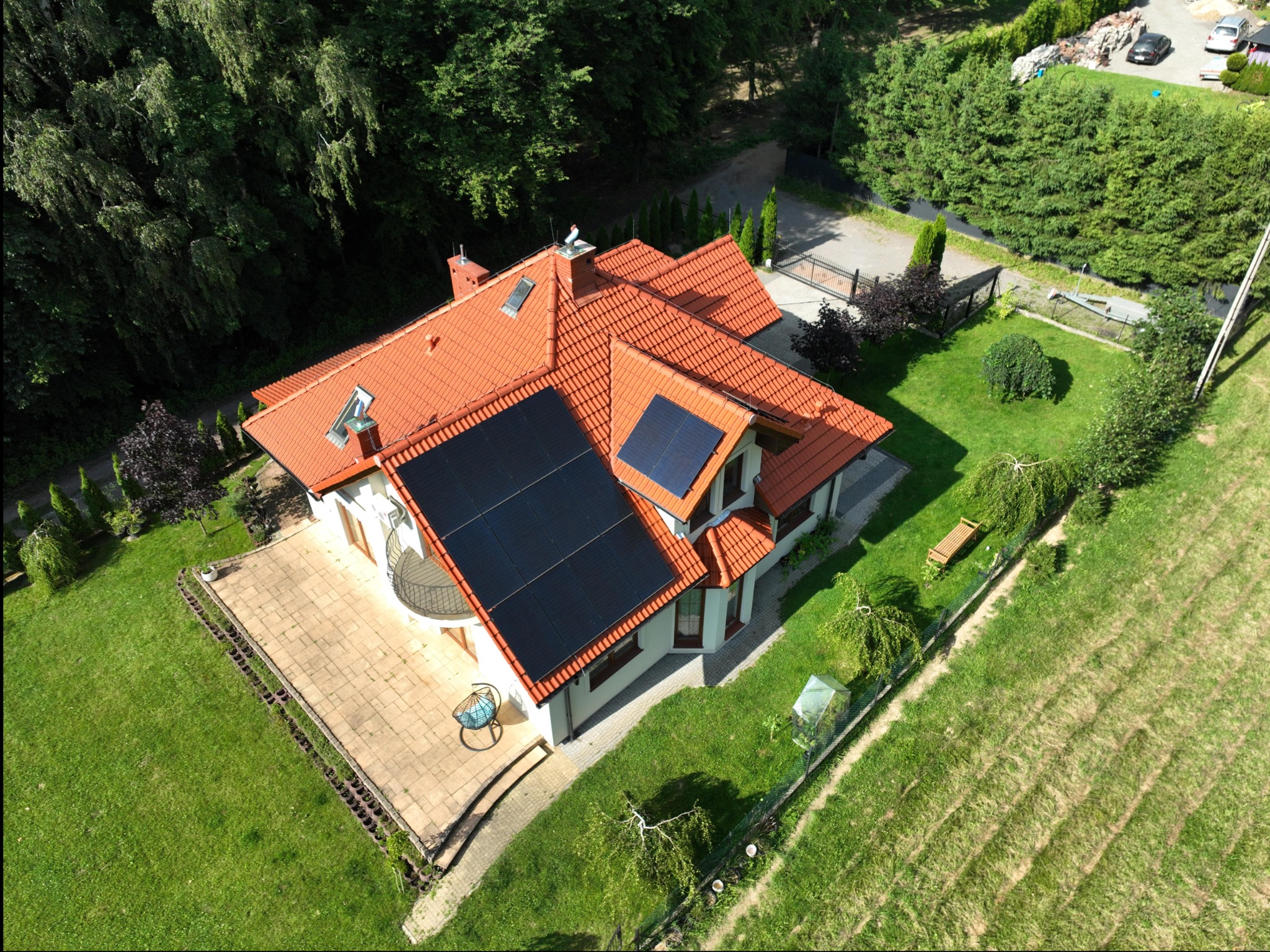 Widok z drona na dom jednorodzinny z pomarańczową dachówką i panelami słonecznymi na dachu, otoczony zadbanym trawnikiem i zielenią.
