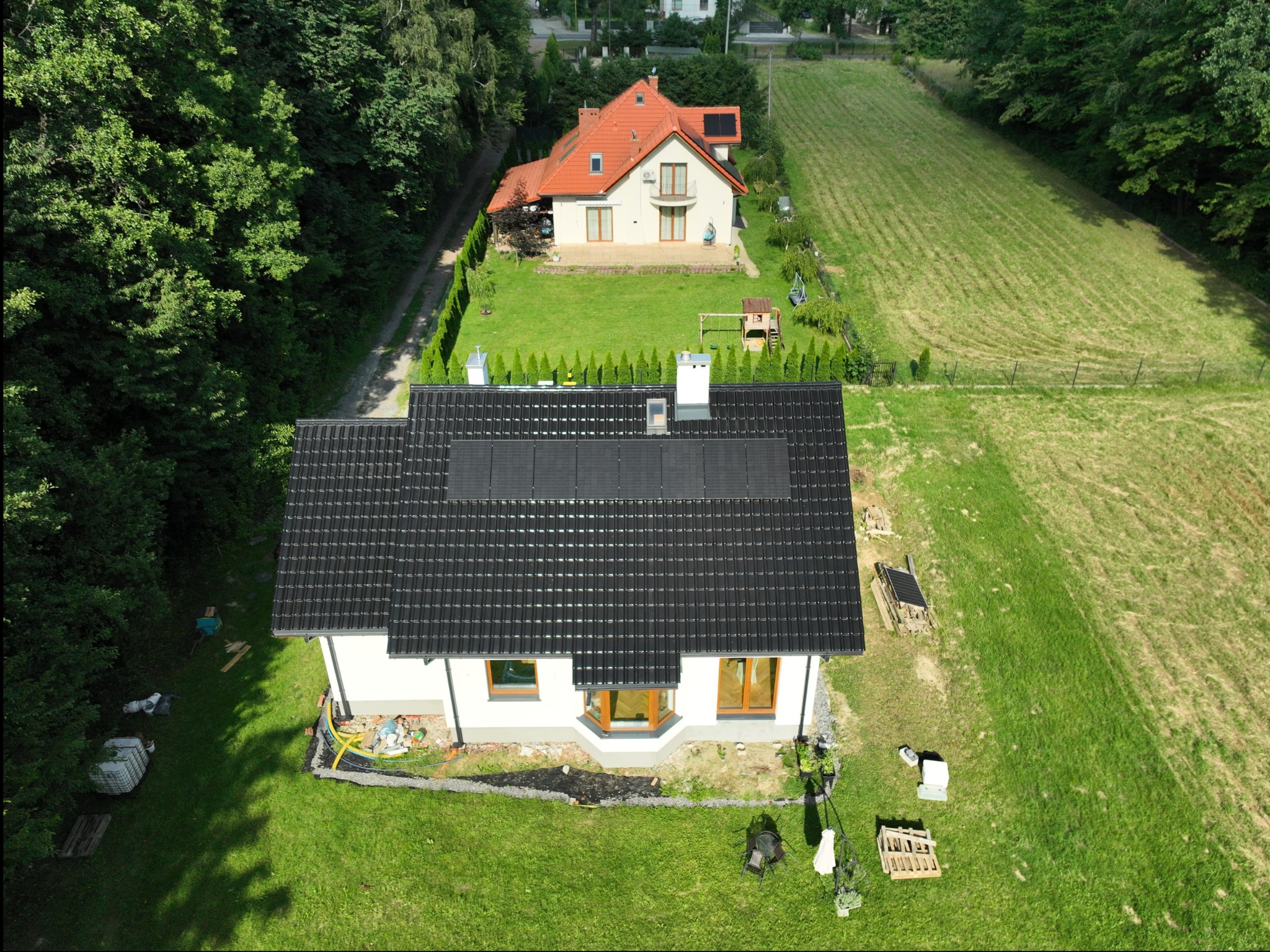 Widok z drona na dom z ciemną dachówką i panelami słonecznymi, w tle drugi dom z pomarańczową dachówką, otoczenie: trawnik, drzewa, ogrodzenie.