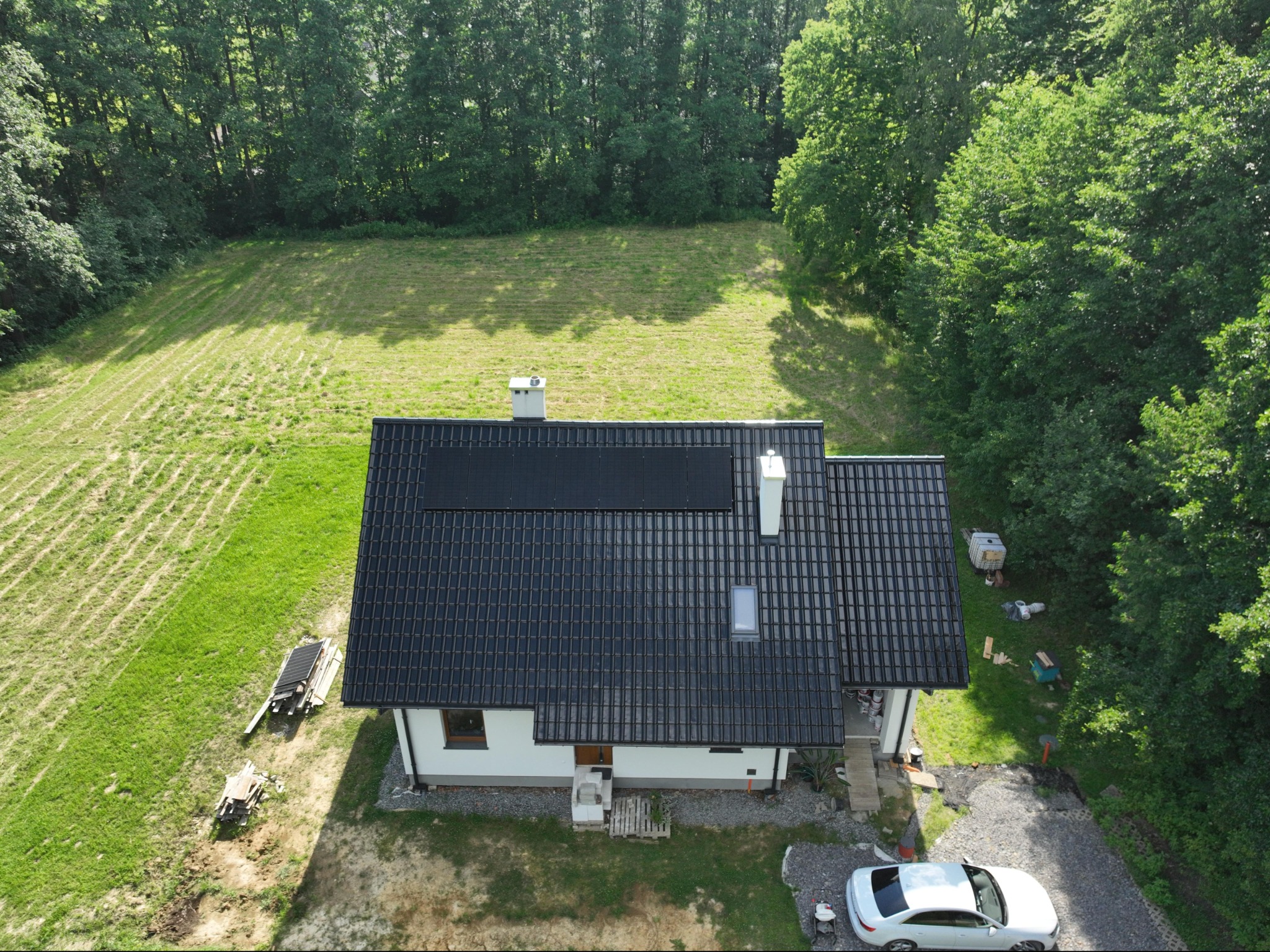 Widok z góry na dom jednorodzinny z ciemną dachówką i zamontowanymi panelami słonecznymi; obok domu zaparkowany biały samochód, a w tle zielony teren z drzewami.