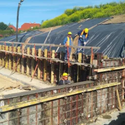 Budowa żelbetowej konstrukcji oporowej: pracownicy w żółtych kaskach układają szalunek z metalowymi wzmocnieniami, widoczny betonowy element i zbocze zabezpieczone folią, w tle zabudowa...