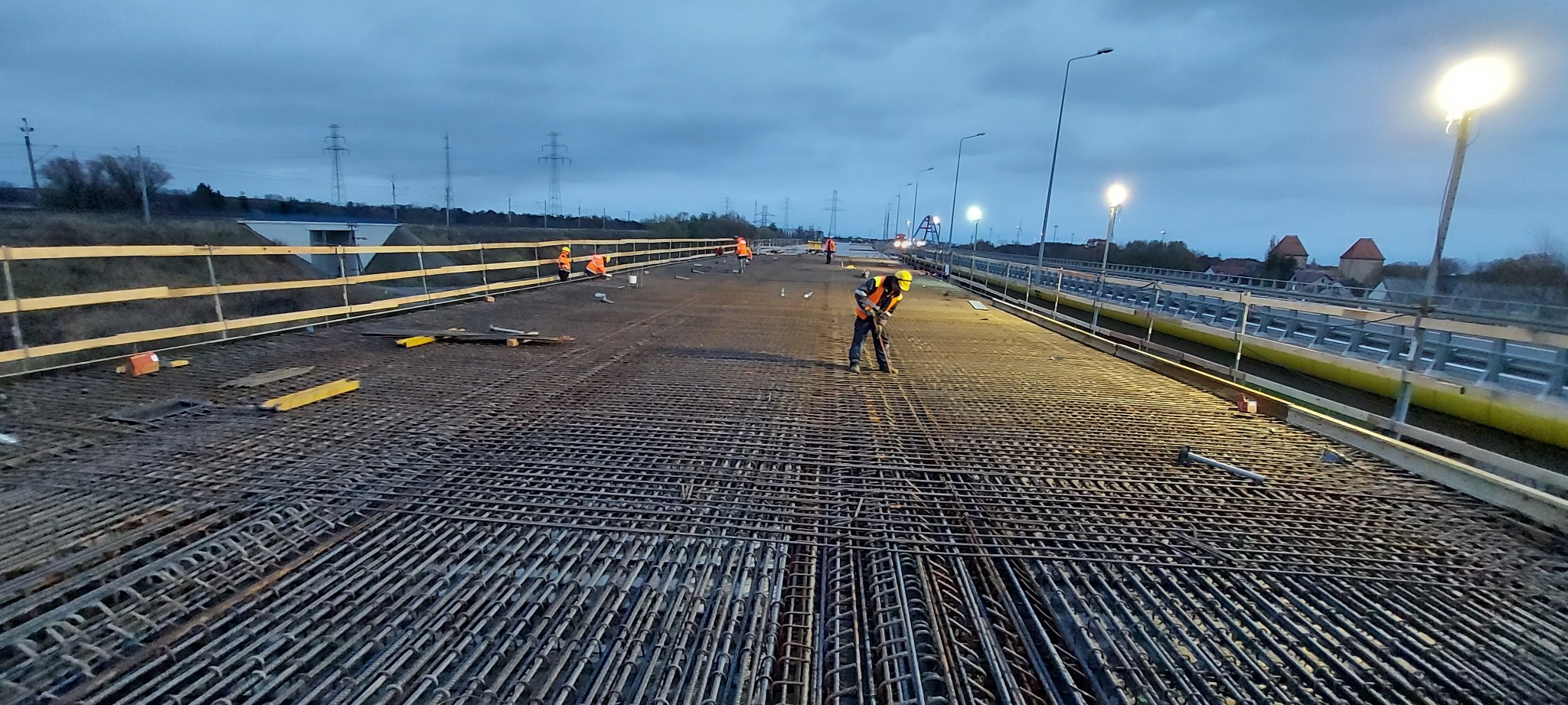 Zbrojenie mostu w trakcie budowy drogi S3 Wolin. Widoczni pracownicy w odblaskowych kamizelkach i kaskach. W tle słupy wysokiego napięcia i oświetlenie.