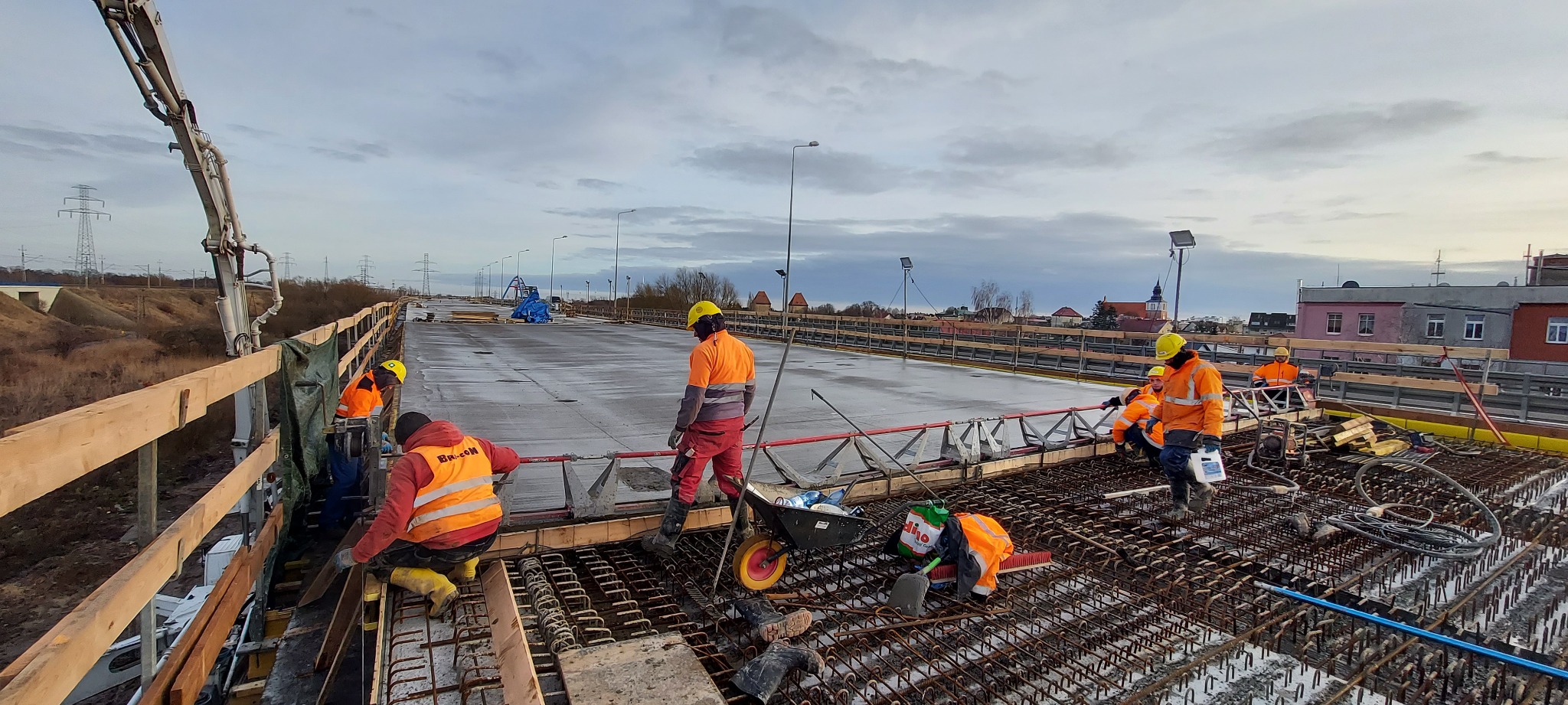 Budowa mostu na trasie S3 Wolin: robotnicy w pomarańczowych strojach układają zbrojenie na betonowej płycie, widoczna pompa do betonu i panorama miasta w tle.