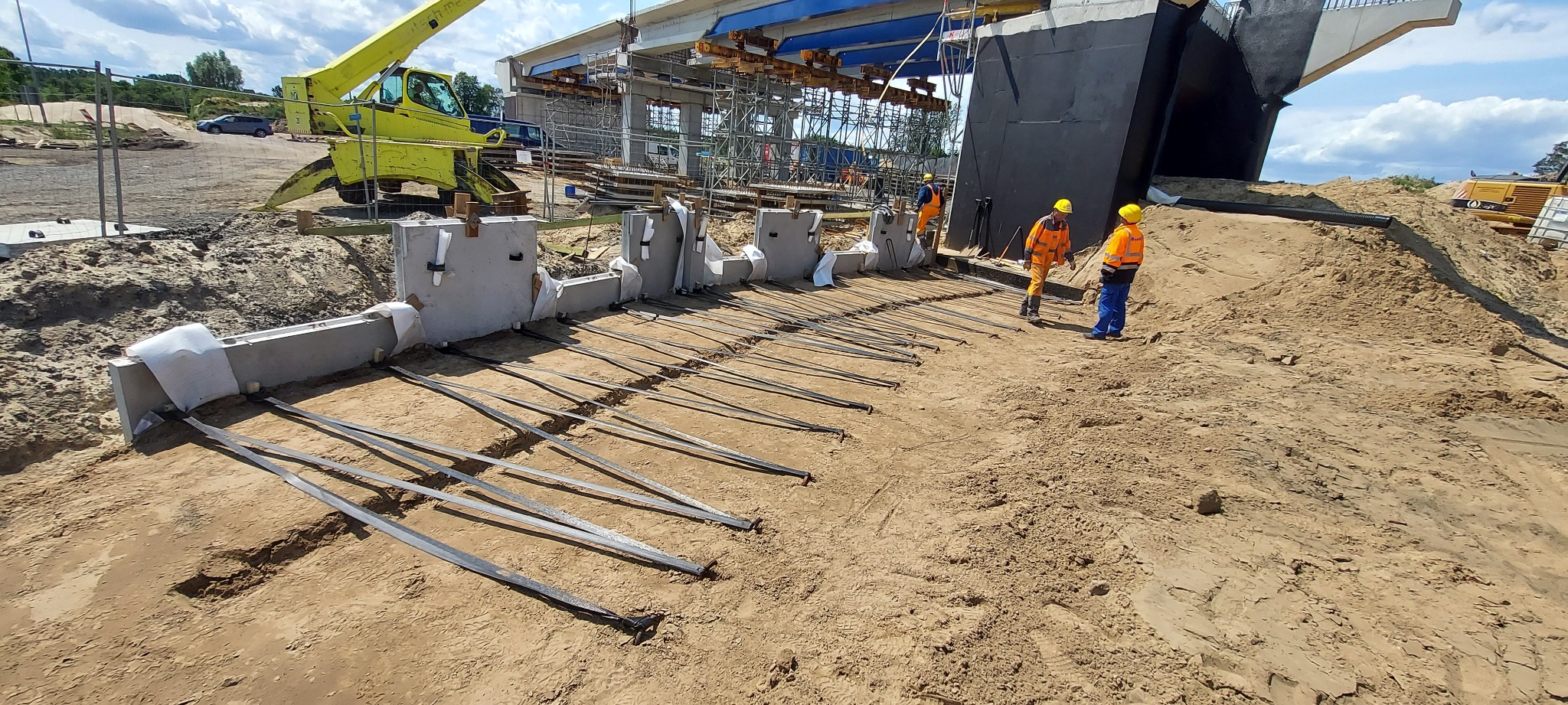 Budowa drogi ekspresowej S3 Wolin: betonowe elementy konstrukcyjne i wystające pręty zbrojeniowe na piaszczystym podłożu. Widoczni pracownicy w odblaskowych ubraniach.