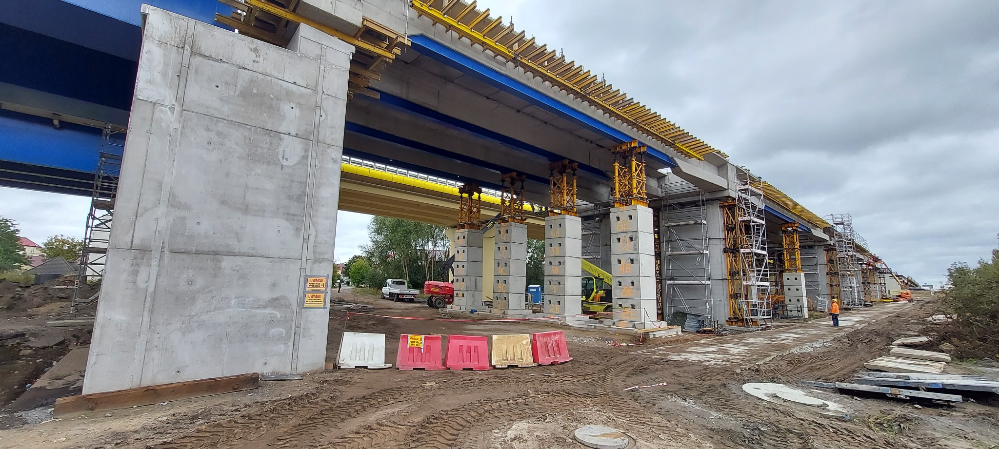 Budowa wiaduktu Wolin S3: betonowe filary, stalowe rusztowania i żółte elementy konstrukcyjne pod pochmurnym niebem. Widoczne bariery drogowe i sprzęt budowlany.