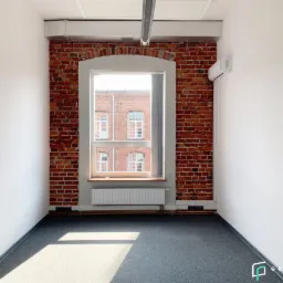 Puste biuro z ceglaną ścianą, dużym oknem z widokiem na budynek z czerwonej cegły, szarą wykładziną i klimatyzacją na ścianie.